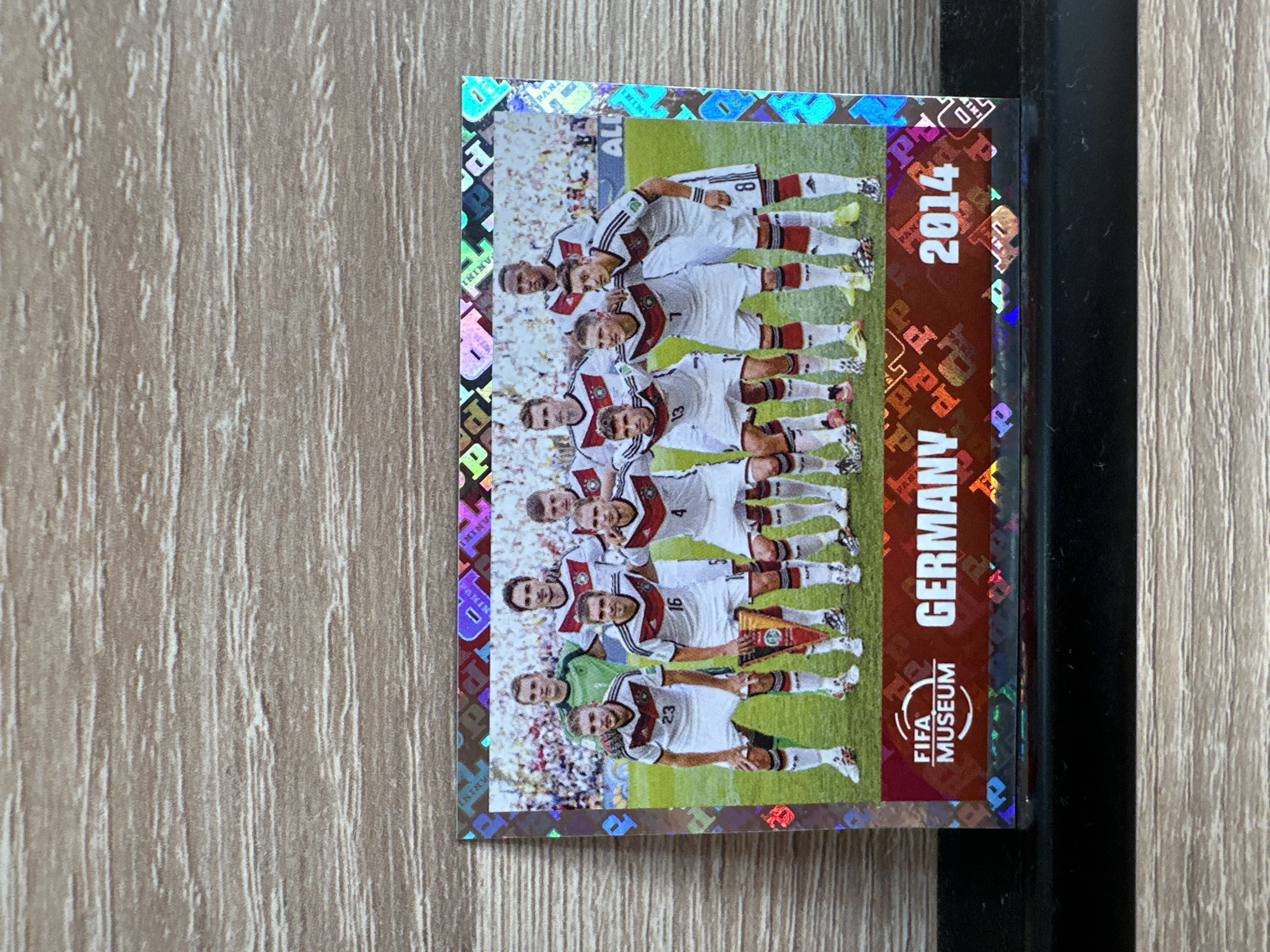 This is a close-up, vertically oriented image of a Panini soccer sticker featuring the Germany national team from the 2014 FIFA World Cup. The sticker has a holographic, diamond-patterned border. The main subject is a team photo of the German players, with eleven players visible, most of whom are in white jerseys with black and red trim, and black shorts. They are posing on a green soccer field with a crowd visible in the background. Some players are standing in two rows, while others are kneeling or sitting. One player, seemingly a goalkeeper, is wearing a green jersey.
Notable details include the year "2014" printed prominently on the right side of the sticker in large white font against a dark red background. Below the year, the words "GERMANY" are vertically oriented in white letters. Further down on the red background, there is a circular logo with the text "FIFA MUSEUM" and a stylized globe. Some of the players have numbers visible on their jerseys, such as "23", "16", "4", "13", and "8". The overall emotion conveyed is one of pride and accomplishment, typical of a team celebrating a victory or a significant tournament. The setting appears to be a professional soccer stadium. The background suggests a sunny day, as indicated by the bright lighting and the presence of a crowd. There is no discernible time of day or weather beyond what is typical for a soccer match. The image is sharp and clear, highlighting the details of the sticker and the players. The sticker itself is in excellent condition, with no visible damage or wear.