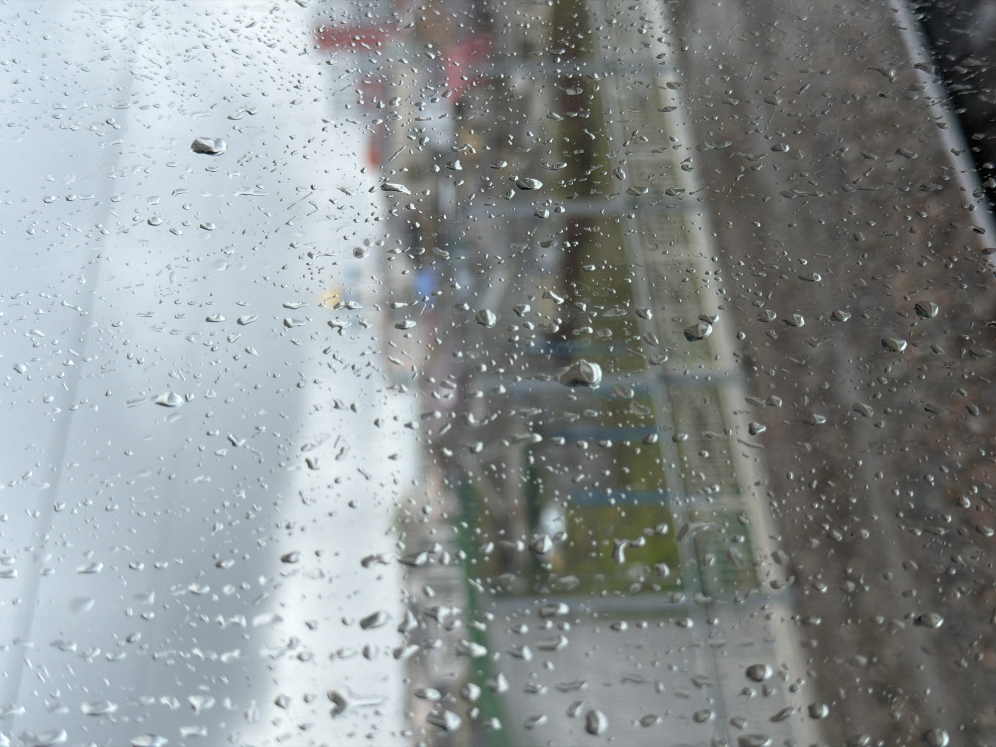 The image shows a view through a rain-covered window. Outside the window appears to be a construction site with industrial equipment and structures, vaguely distinguishable through the water droplets. There are also some hints of greenery visible in the background. The location is purportedly Liski, Russia. No people are visible in the image.