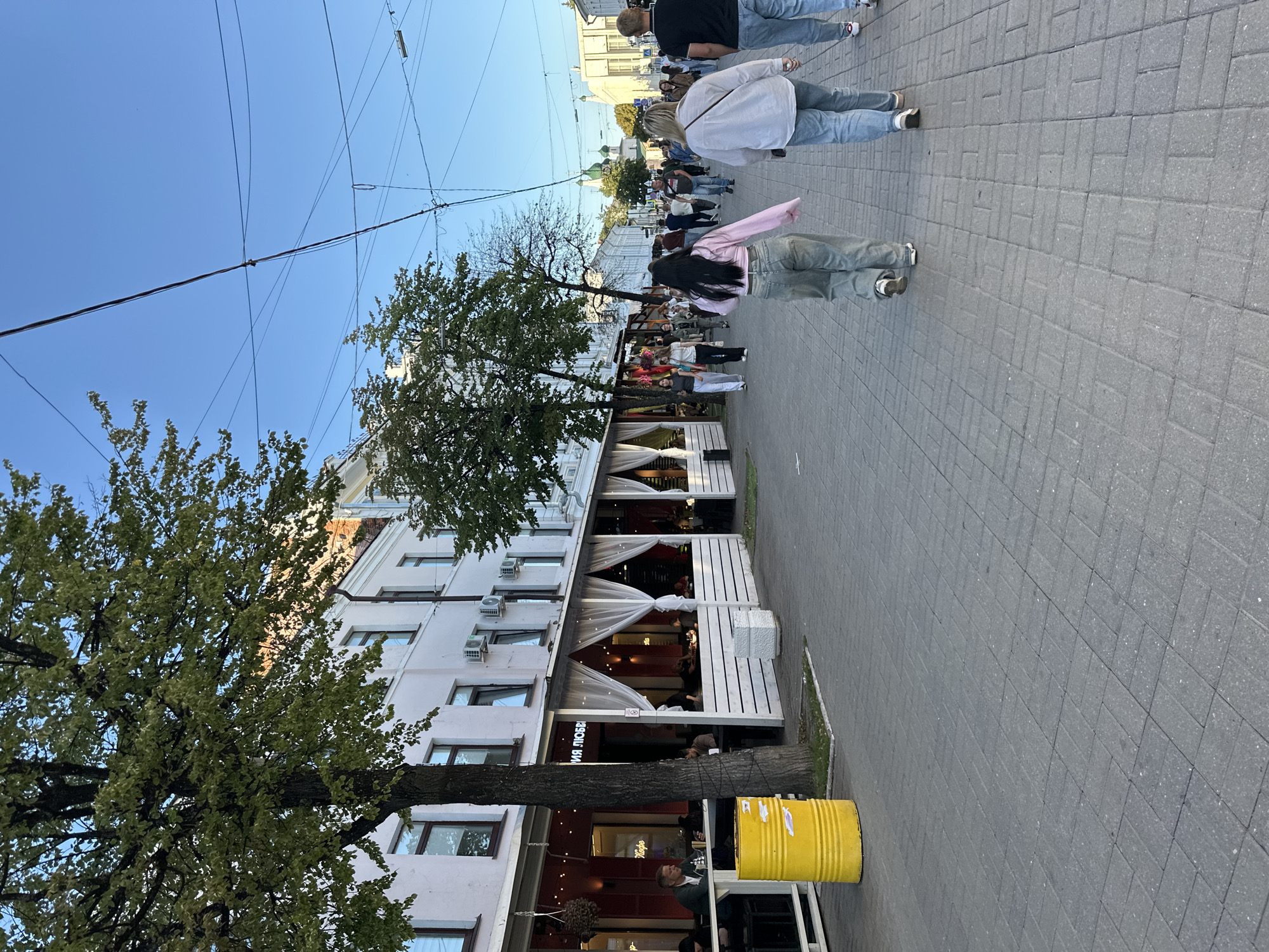 The image depicts a street scene in the city of Feklino, Russia. A pedestrian walkway paved with gray rectangular bricks runs through the center of the frame, lined by buildings and trees. On the left side of the walkway is a restaurant with an outdoor seating area covered by white curtains and string lights. Several people are seated at tables inside and outside the restaurant. A large yellow cylindrical trash bin is positioned next to a tree in front of the restaurant. A group of people, including women and men, are walking away from the camera on the pedestrian walkway. Some of the individuals are dressed in casual clothing, such as jeans and t-shirts. In the background, buildings with white facades and windows are visible, as well as trees and a clear blue sky. Overhead, electrical wires are strung across the sky. The overall impression is of a bustling urban environment with commercial activity and people enjoying a pleasant day.
