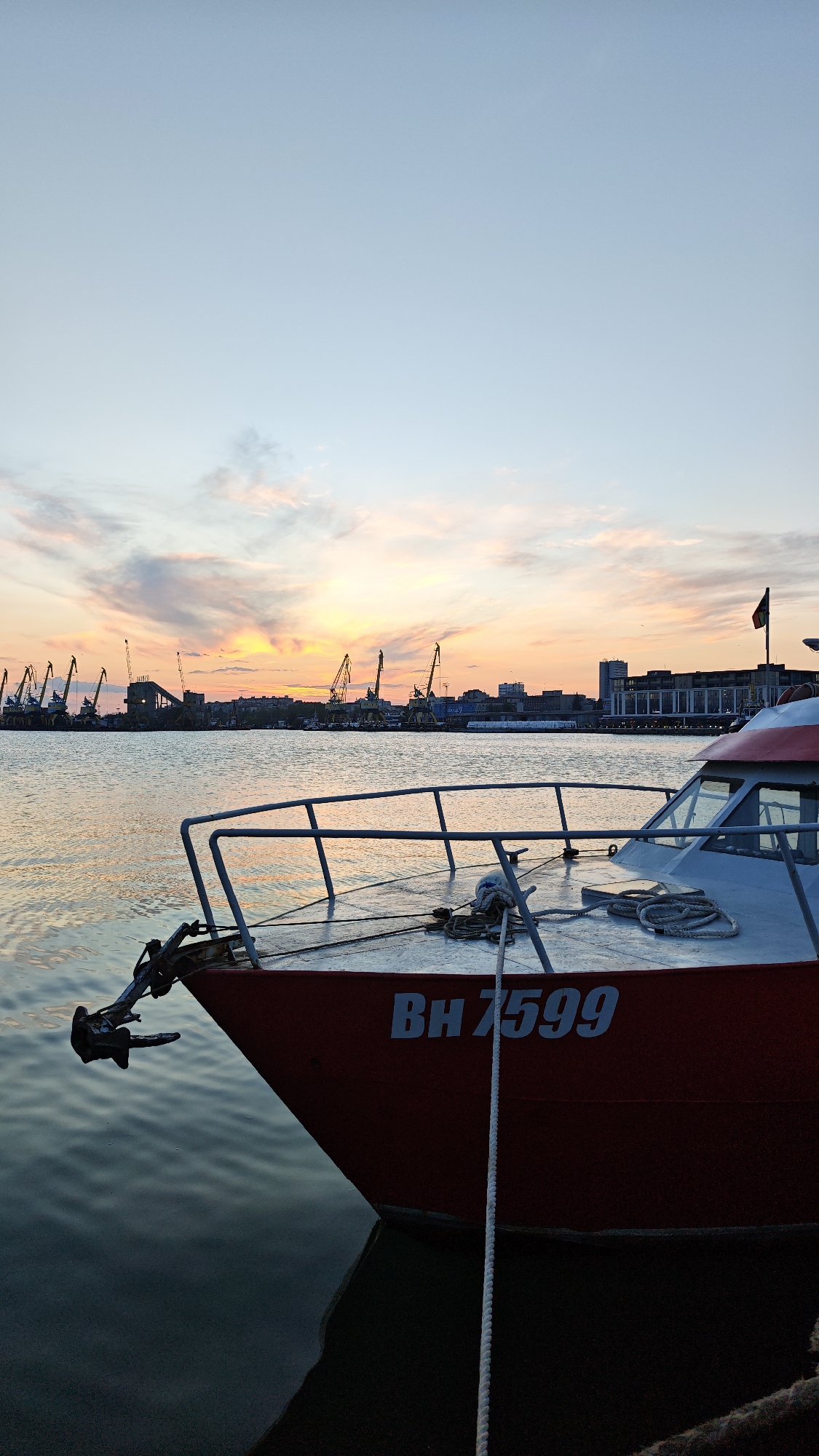 The image depicts a boat docked in the harbor of Burgas, Bulgaria, during the early evening, indicated by the colorful sunset. The boat's hull is primarily red with white accents on the deck and cabin. Visible on the hull is the registration number "BH 7599". A mooring rope hangs vertically down the bow, entering the water. The anchor is visible at the front of the boat.
In the background, across the water, are the silhouettes of industrial port cranes and city buildings. The cranes suggest ongoing port operations. The skyline includes buildings of varying heights, indicating a mixed urban environment. The water is calm, reflecting the colors of the sunset.
