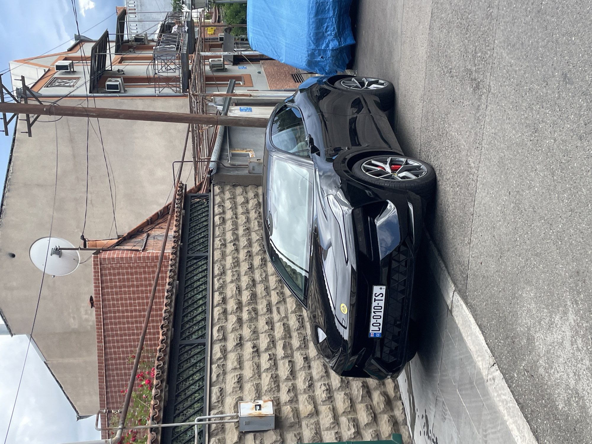 The image depicts a black Lotus Emira coupe parked on a narrow street in Tbilisi, Georgia. The car, displaying a Georgian license plate (LO-010-TS), is parked alongside a stone wall. The background features a cluttered urban environment with buildings displaying varied architectural styles, evident aging, and numerous air conditioning units. Utility lines are visible overhead. A blue tarp covers an object in the background. The street itself is grey asphalt.