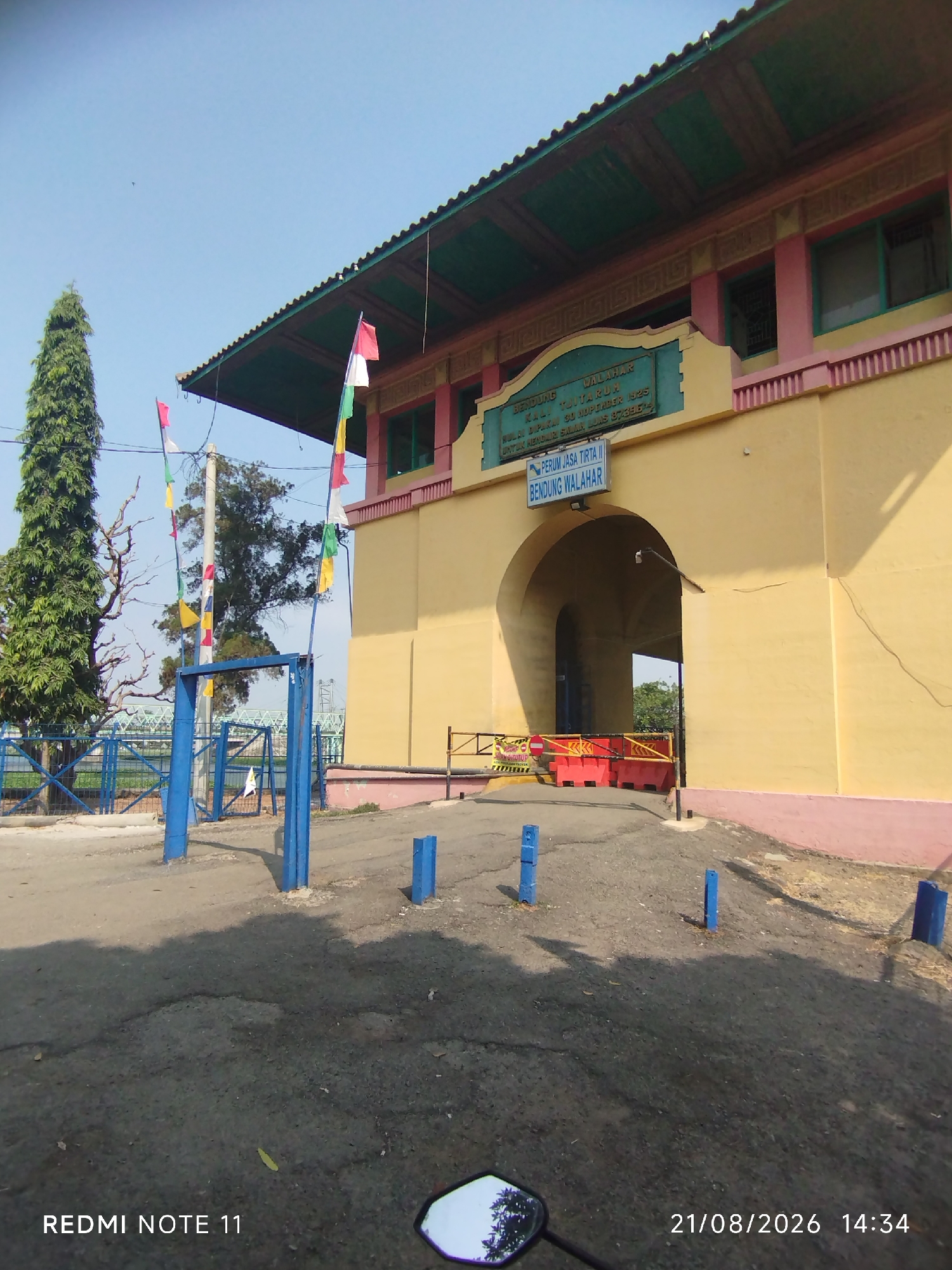 This is a medium-shot, eye-level, outdoor photograph taken on a sunny day at the Bendung Walalahar dam in Karawang, Indonesia. The image features an arched gateway of a yellow building with pink and green accents, topped by a green roof. Flags of red and white, along with colored pennants, are flying from the building's facade and poles. The foreground shows an asphalt road with some cracks and a few blue posts. To the left, a blue metal fence encloses a grassy area with a tall, slender tree. The background reveals a distant bridge, trees, and a clear blue sky. Text on the building identifies it as "PERUM JASA TIRTA II BENDUNG WALAHAR." The bottom left of the image displays "REDMI NOTE 11" and the bottom right shows "21/08/2026 14:34". No people are visible in the frame.