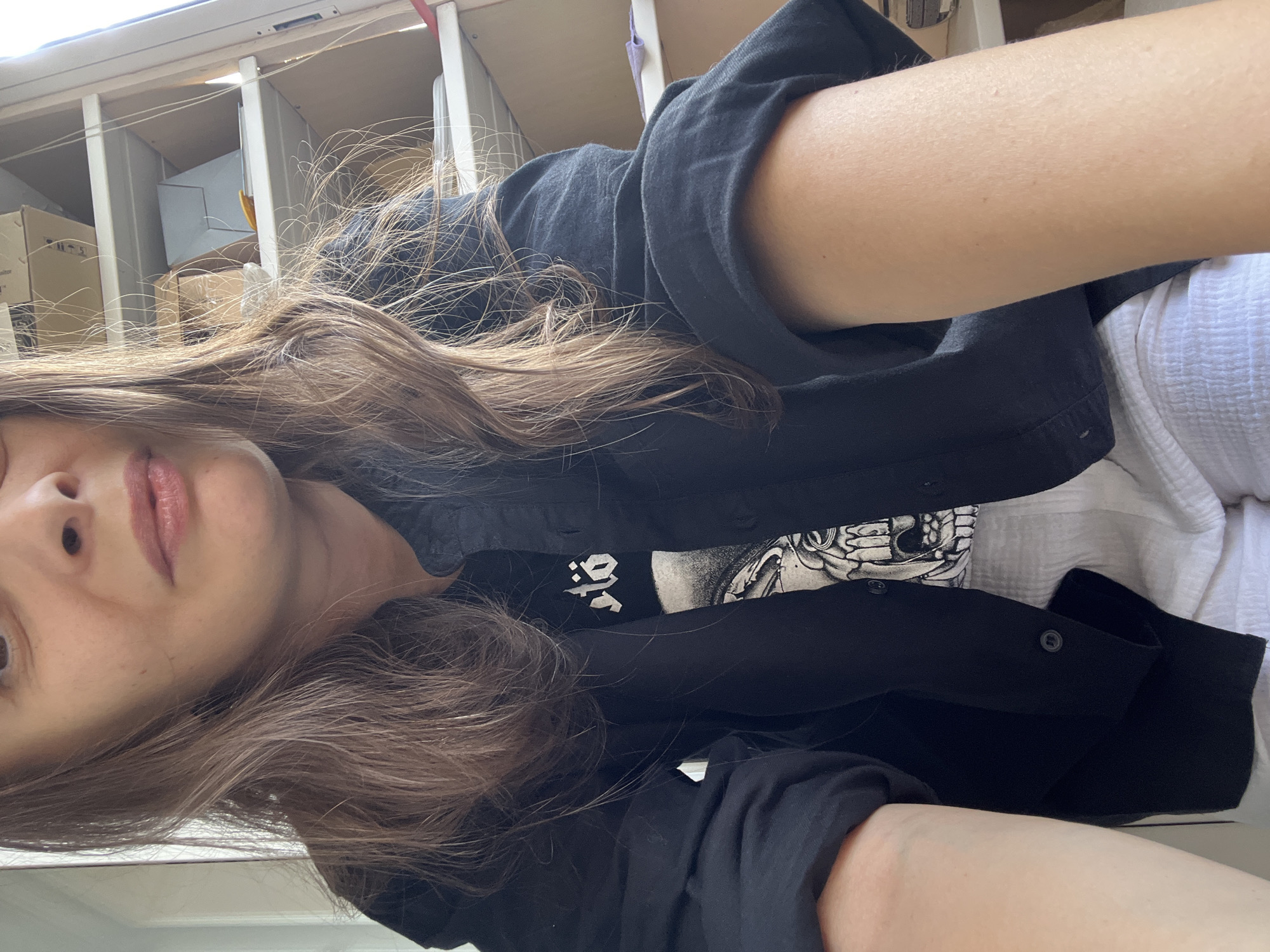 This is a medium-angle, close-up, selfie-style shot of a woman. She has long, brown hair and is wearing a black button-up shirt over a black t-shirt with a graphic design. The woman's face is partially visible, showing her eyes, nose, and mouth. She is also wearing white shorts. The background appears to be an indoor setting with shelves stacked with boxes. The image does not provide any information or context that could be used to determine the location in Tolyatti, Russia.