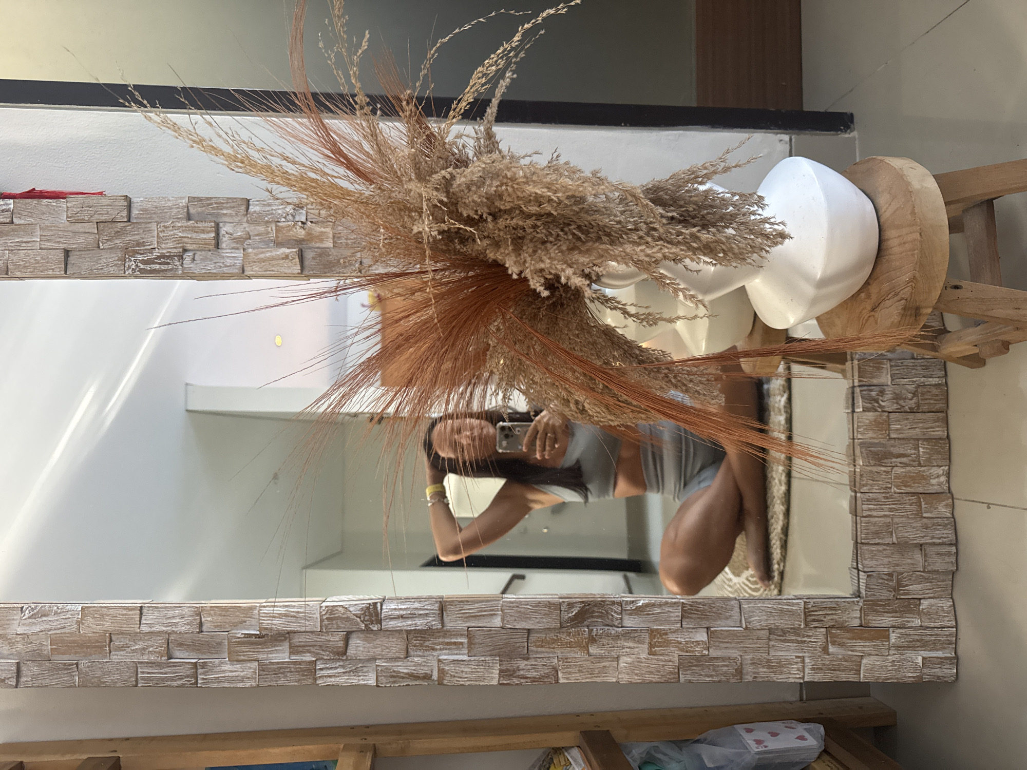 A mirror with a light-colored, brick-patterned frame reflects a woman holding a smartphone, presumably taking a photo in Unknown, Unknown. The mirror also reflects a white object resembling a mannequin head set on a wooden base, and dried floral arrangements. Below the mirror, there is a shelf with various items, including what appears to be a deck of cards. The background includes glimpses of the interior of a building with a neutral color scheme.