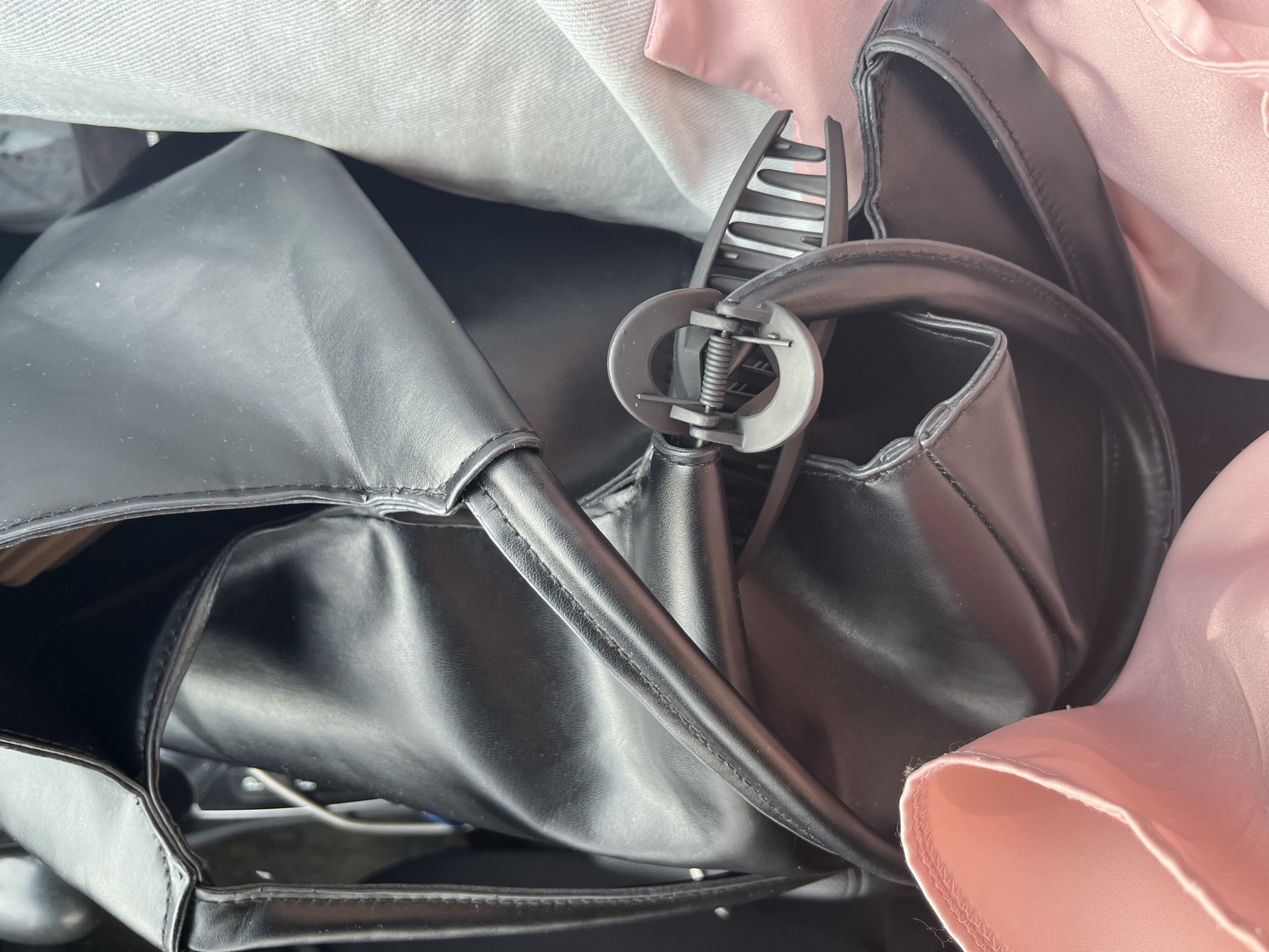The image shows a black handbag with a hair clip. The handbag is made of a shiny material and appears to be partially filled with clothing, including pink and light grey fabric. The scene is cluttered, suggesting it might be inside another bag or a drawer. The lighting is bright and casts shadows, indicating it could be outdoors or near a window. There is no discernible location within the city of Sterlitamak, Russia, as the image provides no contextual clues.