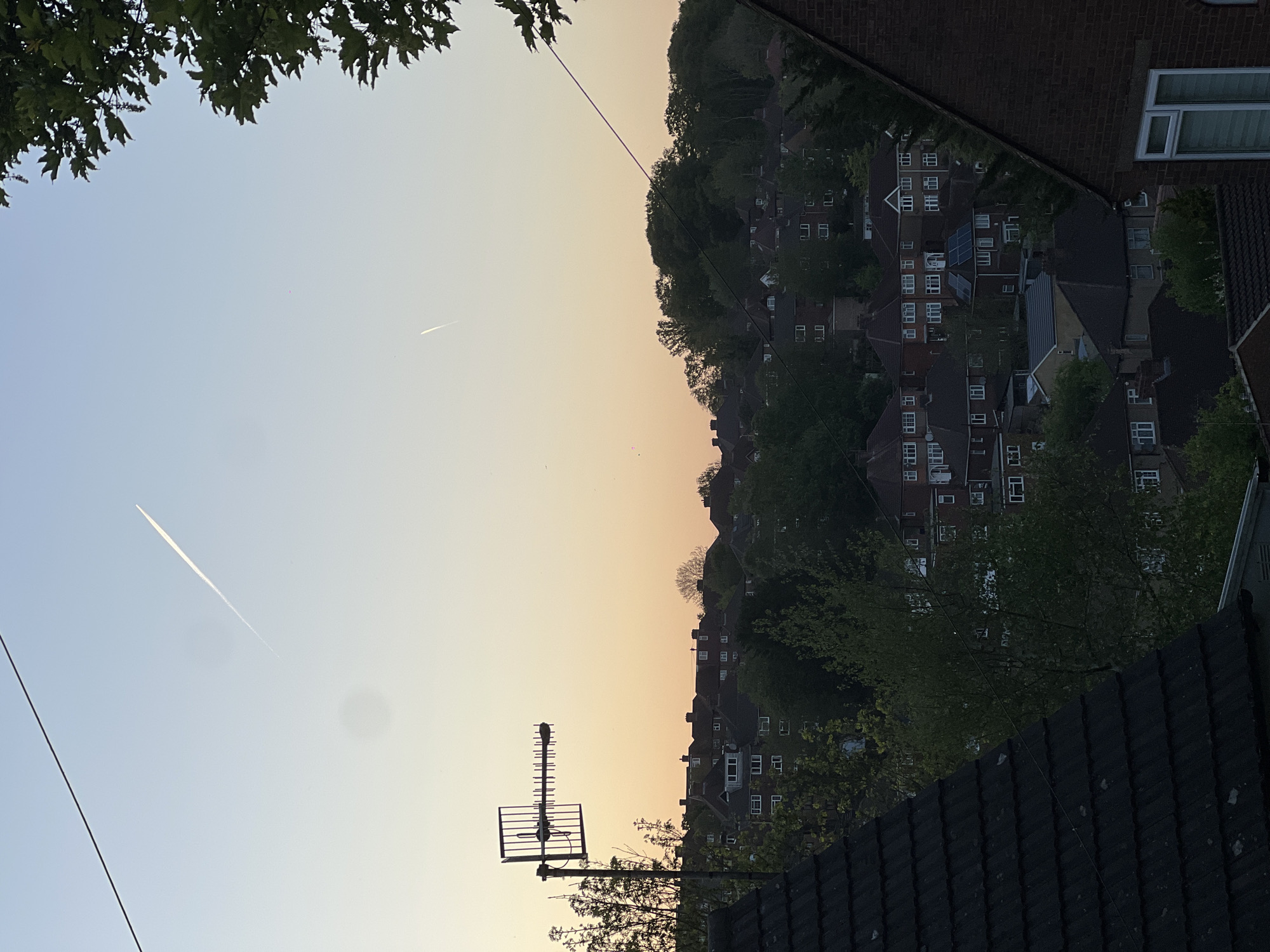 The image displays a panoramic view of a residential area, likely a suburb in High Wycombe, United Kingdom, set against a wide, clear sky. The sky exhibits a gradient from a pale blue overhead to a warmer, peach-orange color closer to the horizon, indicating either sunrise or sunset. Two white contrails from aircraft are visible: one long and distinct in the top-left portion of the sky, and another shorter, fainter one further towards the mid-right horizon.
In the foreground, a dark, tiled roof occupies the bottom-right. To the lower-left, a multi-element television or radio antenna is mounted on a pole, extending upwards from a roof. A thin black wire or cable diagonally crosses the scene, stretching from the upper-left sky down towards the lower-right, passing over houses.
The middle and distant grounds are filled with numerous residential houses, featuring a mix of tiled and flat roofs, white-framed windows, and various architectural styles. Several roofs are equipped with solar panels. A prominent section of a brick building with white windows is visible on the mid-right edge. Verdant green trees are interspersed among the houses and densely cover a distant hillside. A large tree branch with green leaves partially frames the upper-left of the sky. The overall scene conveys a peaceful and serene atmosphere in a typical British suburban landscape. No visible text is present.