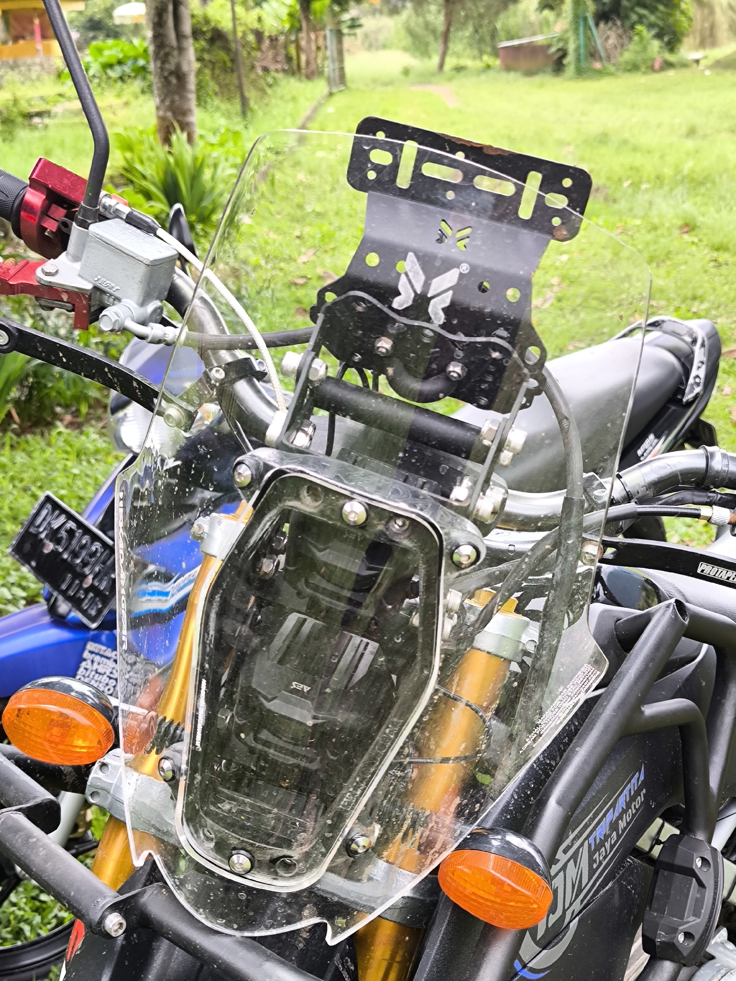 A detailed close-up photograph of the front of a parked motorcycle in Bedugul, Indonesia. The focus is on the motorcycle's transparent windscreen, which is mounted to a black metal frame with visible screws. The handlebars, red brake lever, and other motorcycle components are partially visible. An orange indicator light is also prominently displayed.
In the background, there is lush greenery, including grass and trees. The environment appears to be a natural setting, possibly a park or roadside area. The motorcycle is parked outdoors, suggesting it is either stopped temporarily or in storage.
A partial license plate is visible, labeled DK 5199 FH with dates below. The front of the motorcycle also has text, reading "JM TRIPITIKA Jaya Motor".