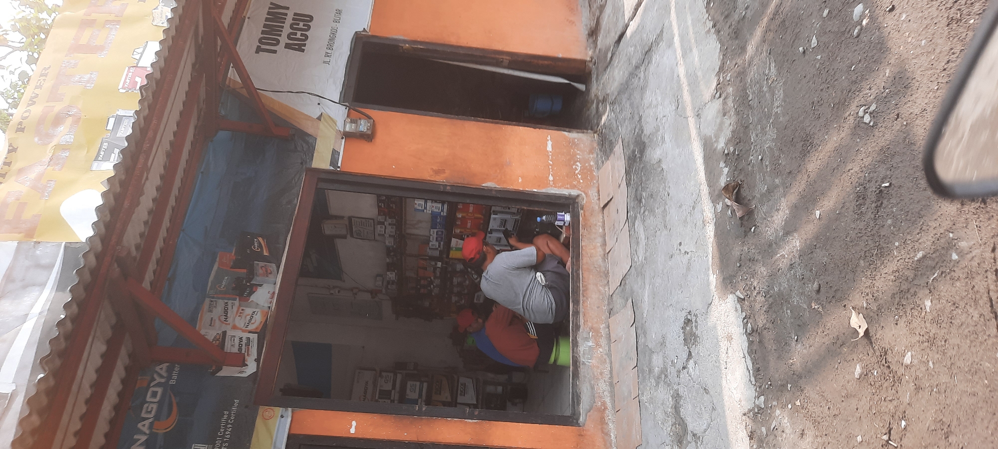 The image captures the storefront of a business named "TOMMY ACCU" located at JL. RY. BRONGKOS - BLITAR in Kesamben, Indonesia. The building has an orange facade. In front of the open doorway, a person wearing a grey t-shirt and shorts is squatting, looking at a mobile phone. To their left, another person in a red shirt and a third person in a blue shirt are also seated. The interior of the shop displays various car batteries, some in their packaging. Above the entrance, there's a corrugated metal roof with red trim. A large yellow banner with "FASTER POWER" written on it is visible on the upper part of the building, along with another banner featuring the "NAGOYA Battery" logo. The ground in front of the shop is a mixture of concrete and dirt, with several pieces of cardboard laid out on the ground. Shadows from nearby trees are cast on the ground. A bottle of water sits on the threshold.