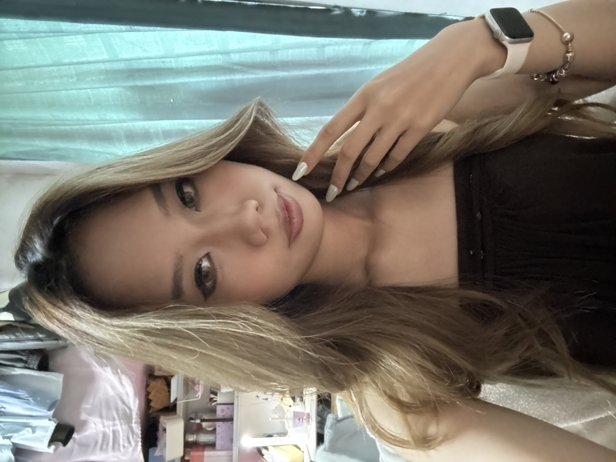 A young woman poses indoors with light brown and blonde hair, wearing a black strapless top. She has light skin, brown eyes, and wears lipstick. Her right hand is raised near her face, adorned with polished nails. Her left wrist features a smartwatch and beaded bracelet. The background includes a blue curtain and a cluttered shelf with various objects. The location and city of Nan'an District, China are not discernible from the image.