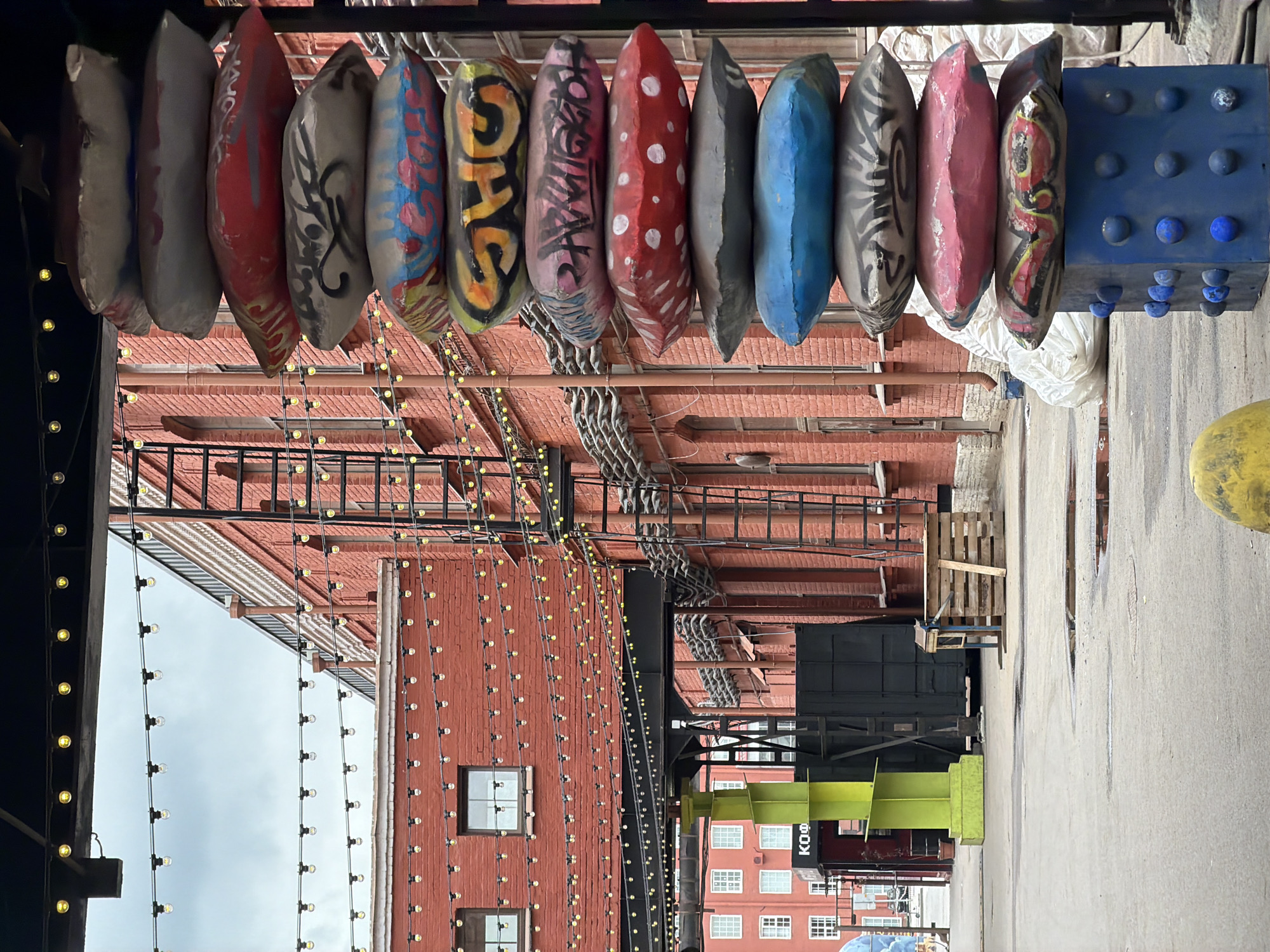 The image depicts a narrow street in Saint Petersburg, Russia, likely an alleyway within an older industrial or commercial district. Strings of small, round lights are strung overhead, creating a festive or decorative atmosphere. A brick building rises on one side, featuring multiple windows and fire escapes. On the opposite side, a smaller, lighter-colored building is visible.
A row of painted objects, possibly canoe or kayak-shaped, hangs from the top of the frame. They are decorated with various colors, patterns, and graffiti-style lettering. Below them are various pieces of industrial equipment, including metal framing and what looks like a large wooden pallet. The street surface is made of concrete. A blue structure adorned with protruding blue spheres is positioned above the street.