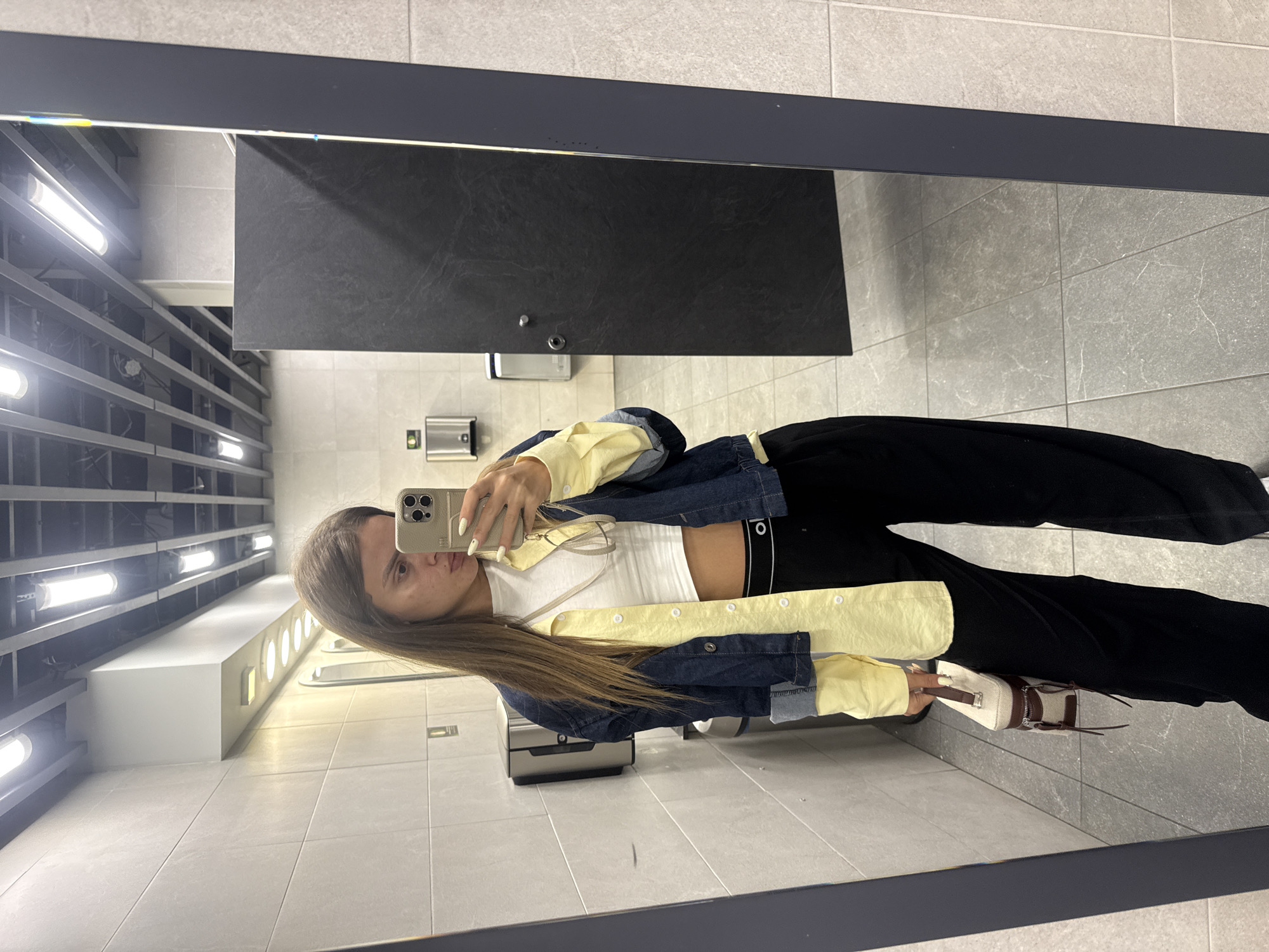 The image shows a young woman taking a selfie in a brightly lit, modern public restroom, likely in a commercial or transportation hub. The woman is standing in front of a large mirror, wearing a white crop top, black wide-leg pants, and a denim jacket layered over a yellow collared shirt. She is holding a small, light-colored handbag. The restroom features light-colored tiled walls and floors, with visible ceiling lights and ventilation. A dark-colored stall door is visible to the right of the woman, and stainless steel dispensers are mounted on the wall. The overall impression is one of a clean, functional, and contemporary public space. The location in Sofia, Bulgaria, is not discernible from the visual information provided.