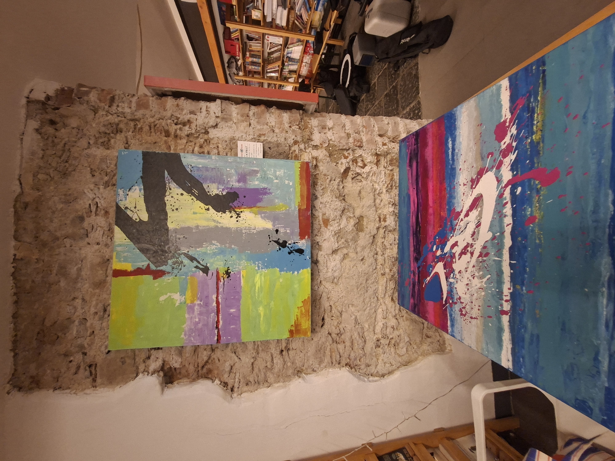 The image shows two abstract paintings hanging on a distressed brick wall. The first painting, on the left, features a gray and black shape on a background of green, blue, and purple stripes. The second painting, on the right, features white paint splatters on a background of blue, pink, and white stripes. The wall is rough and has visible cracks and crumbling plaster near the bottom. A bookcase filled with books and a black bag are visible in the background, above the wall. Based on the available information, it is not possible to confirm if this is in Messina, Italy.