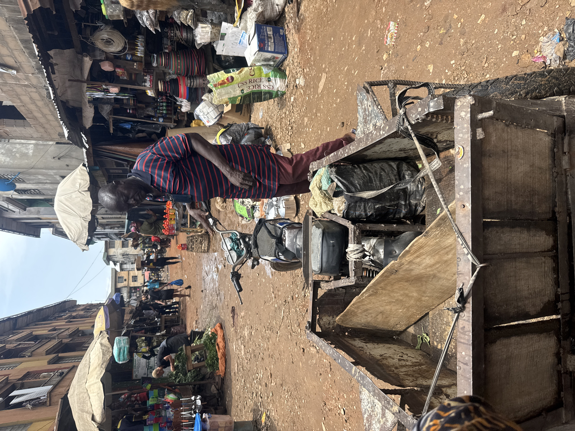 The image depicts a bustling street scene in Onitsha, Nigeria. An elderly man wearing a striped shirt and maroon pants is positioned near a modified motorcycle with an attached cart. The cart is filled with black plastic bags and what appears to be miscellaneous items. The man appears to be interacting with the contents of the cart. In the background, numerous people are walking along a dirt road, and there are stalls and vendors selling various goods. The environment suggests a marketplace or commercial area, with buildings and awnings visible in the background.