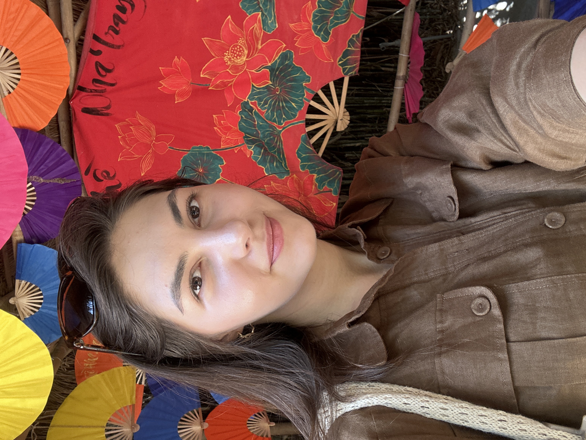 A close-up selfie of a young Caucasian woman with long, dark brown hair and light brown eyes is taken outdoors. She is wearing a brown button-up shirt with two chest pockets and is carrying a woven bag over her left shoulder. She has on small, gold hoop earrings and sunglasses pushed up onto her hair. The background features a collection of colorful paper fans arranged on a bamboo structure. A large red fan with painted lotus flowers and Vietnamese script is prominently displayed behind her. The scene is likely in a market or cultural display area within the city of Nha Trang, Vietnam, as suggested by the Vietnamese script on the fan.