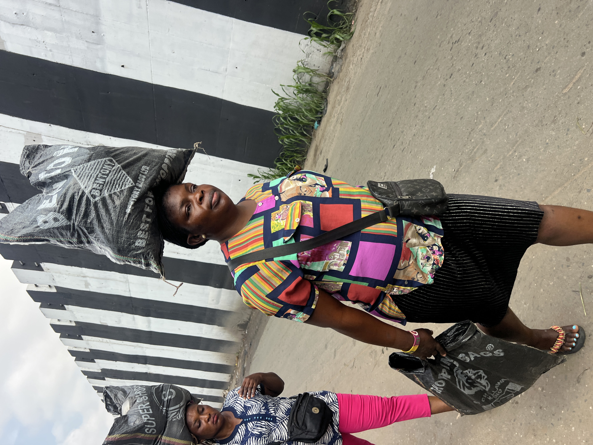 Two women are walking along a road in Nkpor, Nigeria. The woman in the foreground, on the right, is carrying a large black bag on her head. She is wearing a colorful, patterned shirt and a black striped skirt. She has a black crossbody bag and is holding another black plastic bag in her left hand. The woman in the background, on the left, is also carrying a black bag on her head. She is wearing a blue and white patterned shirt and bright pink pants. She has a black fanny pack around her waist. They are walking next to a concrete wall with alternating black and white stripes. Green plants are growing along the base of the wall. The road appears to be paved.