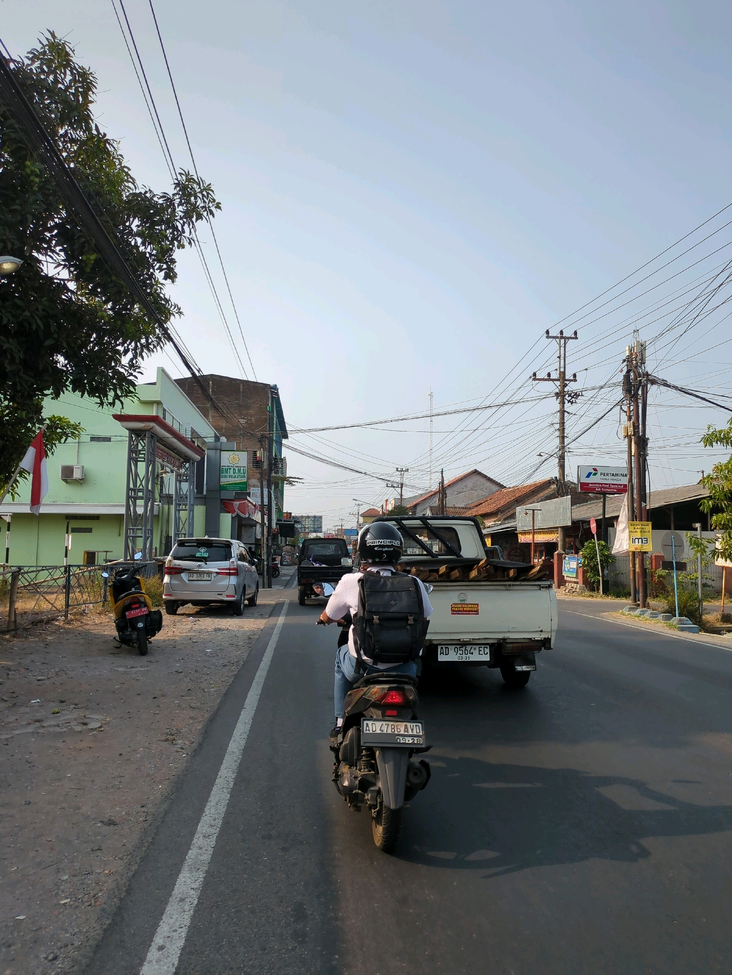 The image depicts a street scene in Desa Ngesrep, Indonesia, under a clear sky. A person on a motorcycle, wearing a helmet and a backpack, is traveling on the road, with a white pickup truck ahead of them. The road is paved and has a white lane marker. On the left side of the road, there are trees, a light green building with an archway, and several parked motorcycles. The right side of the road features more buildings, utility poles with numerous wires, and signs, including one for "Pertamina." The overall impression is of a typical day in a bustling Indonesian town, with traffic and commercial activity.