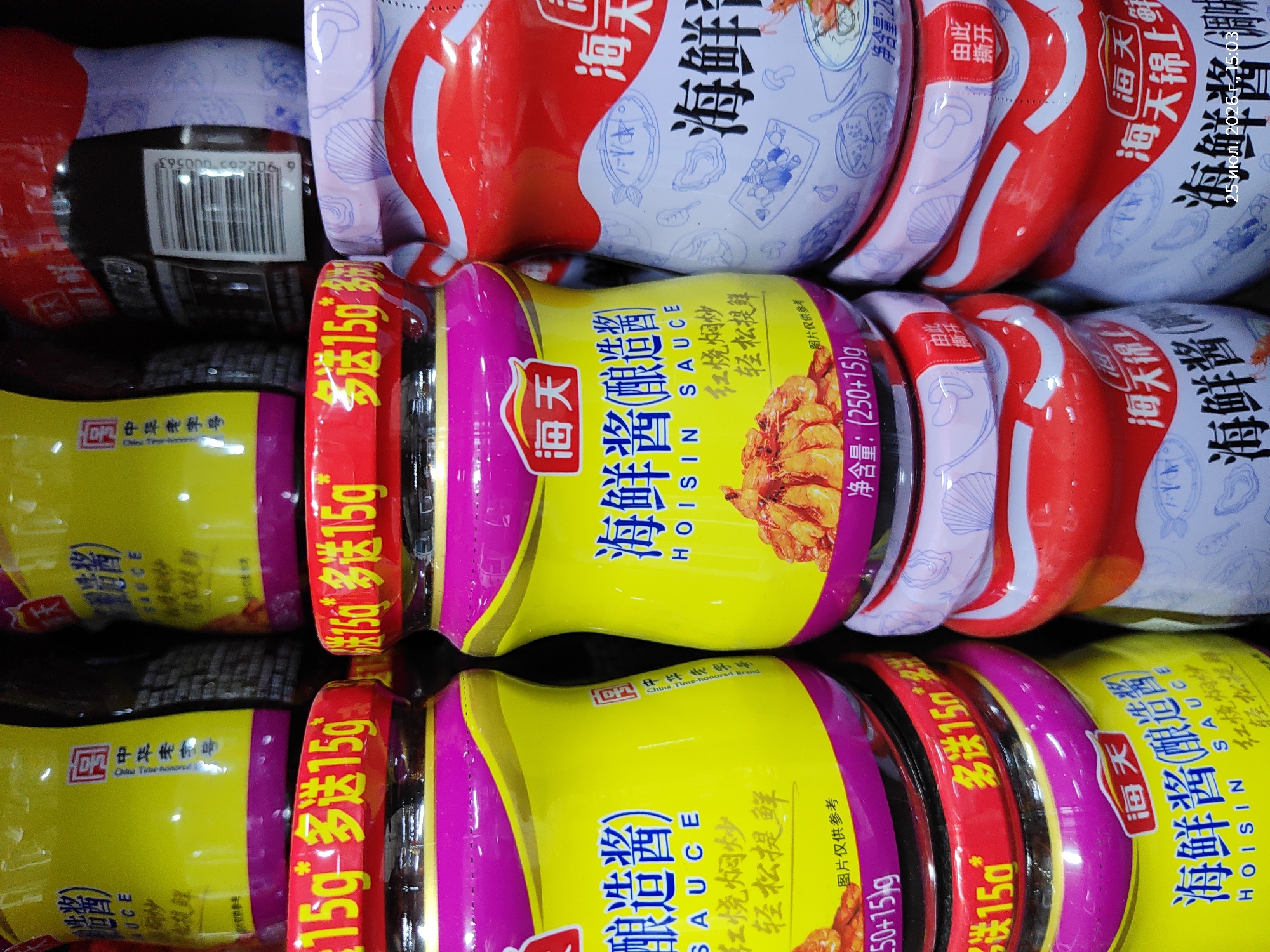 The image shows a close-up view of several stacked jars of Hoisin sauce. The jars are predominantly yellow and red, with prominent Chinese characters and branding. Some jars feature a red lid with yellow text that says "多送15g" (meaning "extra 15g"). The overall scene appears to be a retail environment, possibly a supermarket aisle, with the products arranged for display. The lighting suggests an indoor setting, and the focus is on the product labels. There are no people or discernible elements of the city of Astana, Kazakhstan present in the image.