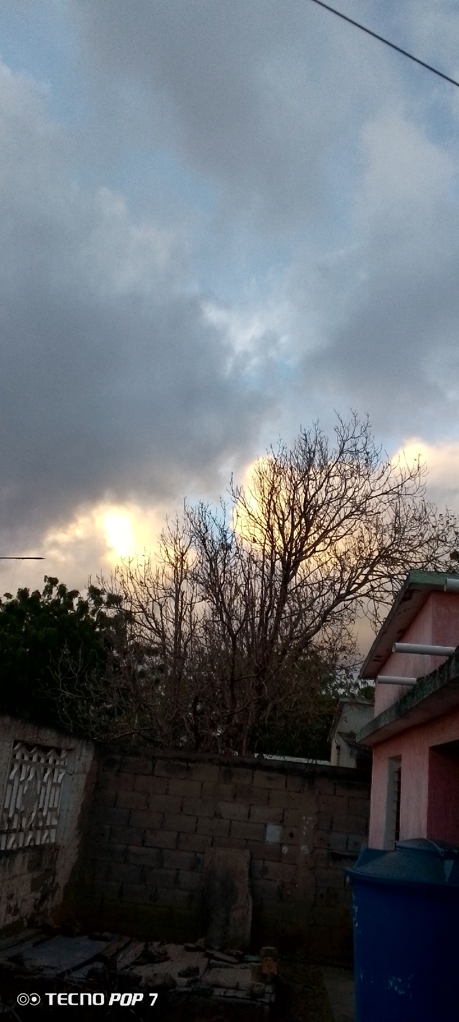 The image depicts a cloudy sky with patches of sunlight breaking through, illuminating the branches of a bare tree. In the foreground, a brick wall runs horizontally across the frame, with a small structure to the left featuring a barred window. To the right, a portion of a pink building with a sloped roof is visible, along with a large blue water tank. The scene appears to be an outdoor setting within a residential area, possibly in Altagracia, Venezuela. The overall lighting suggests either early morning or late afternoon. No people are present in the image.