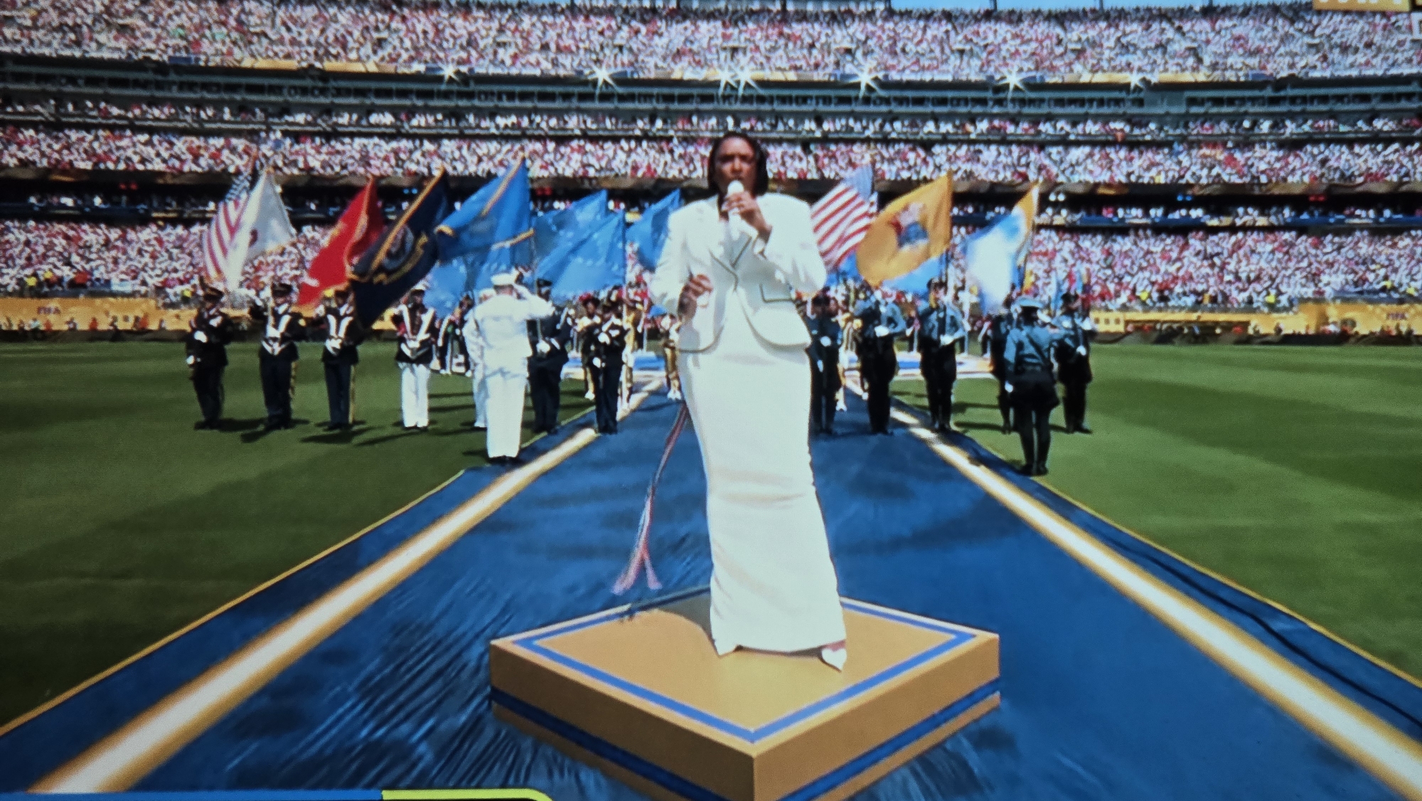The image depicts a large outdoor stadium filled with spectators, indicating a sporting event or a formal ceremony. In the foreground, a woman in a white suit and matching long skirt stands on a raised golden platform, holding a microphone and singing. She is surrounded by a blue carpeted walkway with yellow trim. To her left and right, a group of individuals in formal military-style uniforms are marching, carrying various flags, including the American flag and other national flags. The background is dominated by the stands filled with a diverse crowd. The scene is set on a green grassy field, consistent with a stadium environment. The overall impression is one of a grand performance or national anthem rendition during a significant public event.