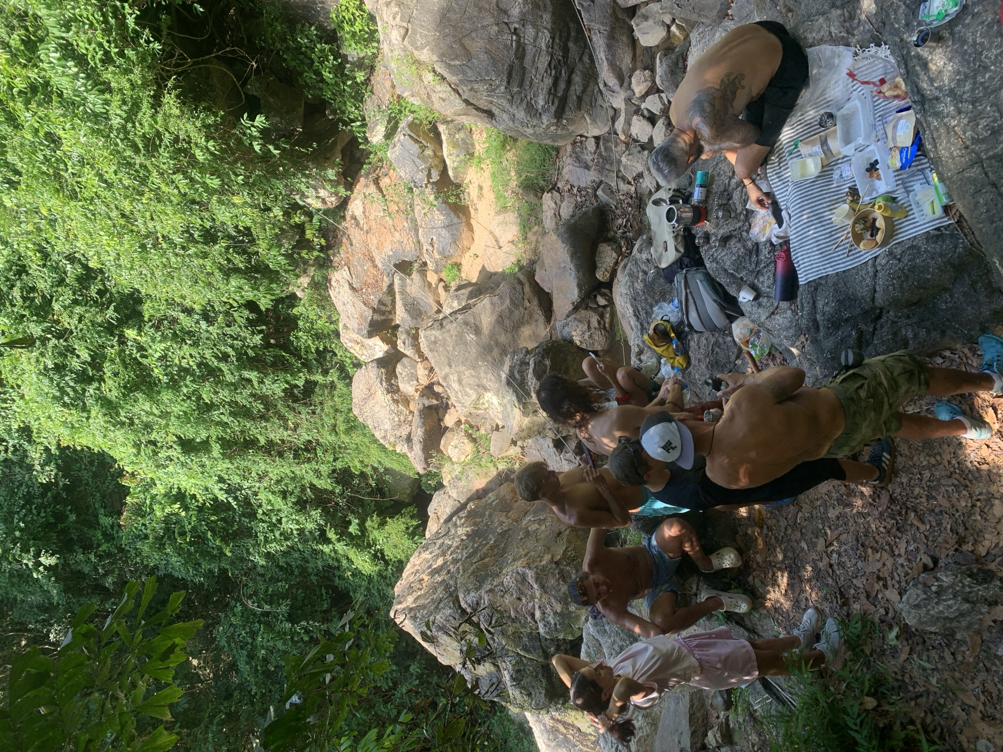 A group of seven people, six male and one female, are gathered on a rocky embankment near a waterfall in a lush, green jungle environment. The individuals are dressed casually, with several of the males shirtless. They appear to be having a picnic, with food and drinks laid out on a striped blanket on a large rock. There are bags, a thermos, and various containers of food visible. The people are interacting with each other and with the food, suggesting a relaxed, social gathering. The rocky terrain and dense foliage indicate an outdoor, natural setting, likely in a remote or park-like area. The presence of a waterfall suggests a water source nearby. The time of day appears to be daytime, with sunlight filtering through the trees, creating shadows on the ground.