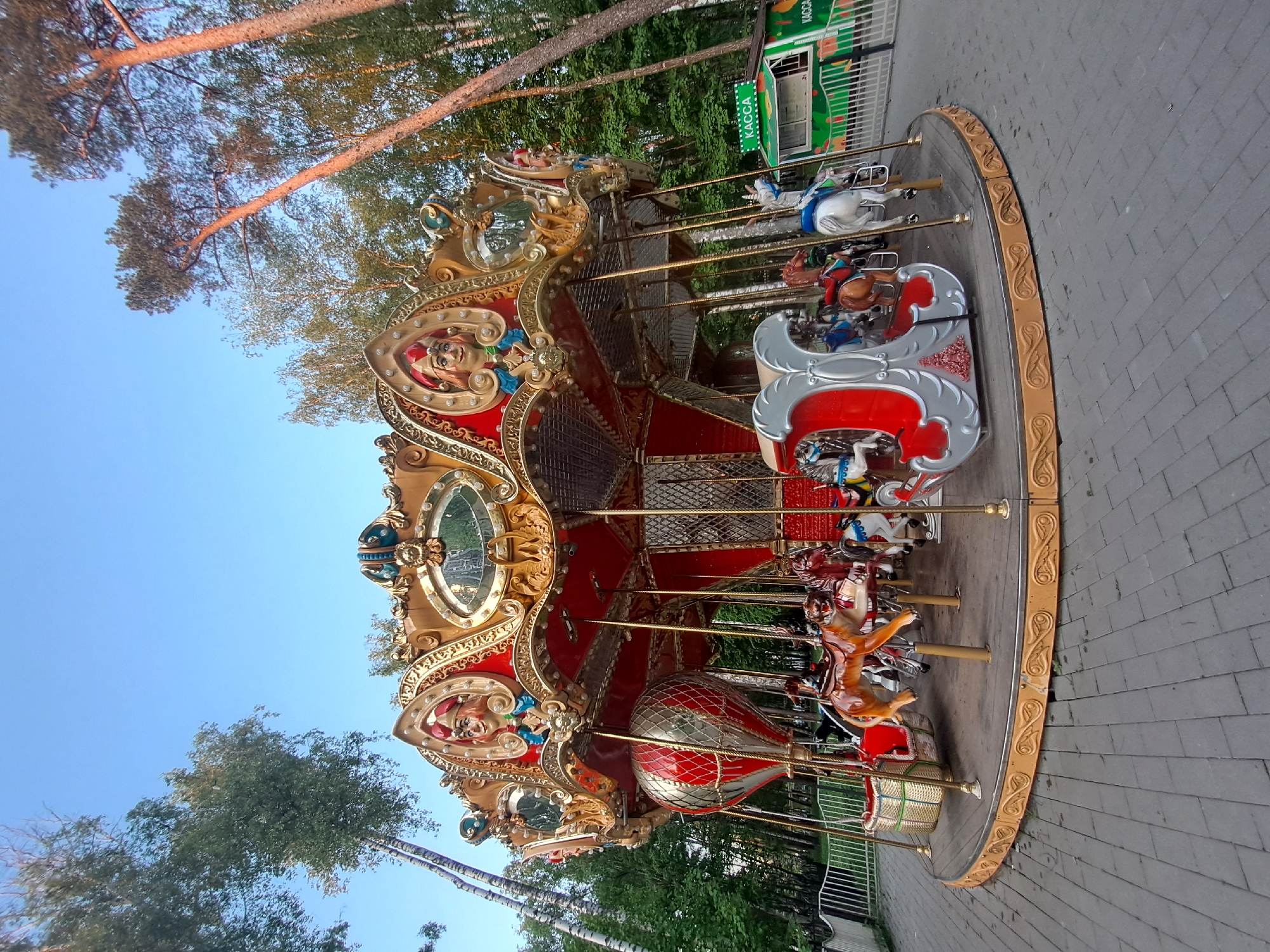 The image shows a carousel in the city of Noginsk, Russia. The carousel is multi-tiered, with ornately decorated horses and other ride vehicles, including an air balloon and a sleigh, positioned around a central pole. The carousel is colored in red, gold, and silver. A ticket booth is visible in the background. Tall trees surround the carousel, and the sky is a clear blue. The ground around the carousel is paved with grey bricks.