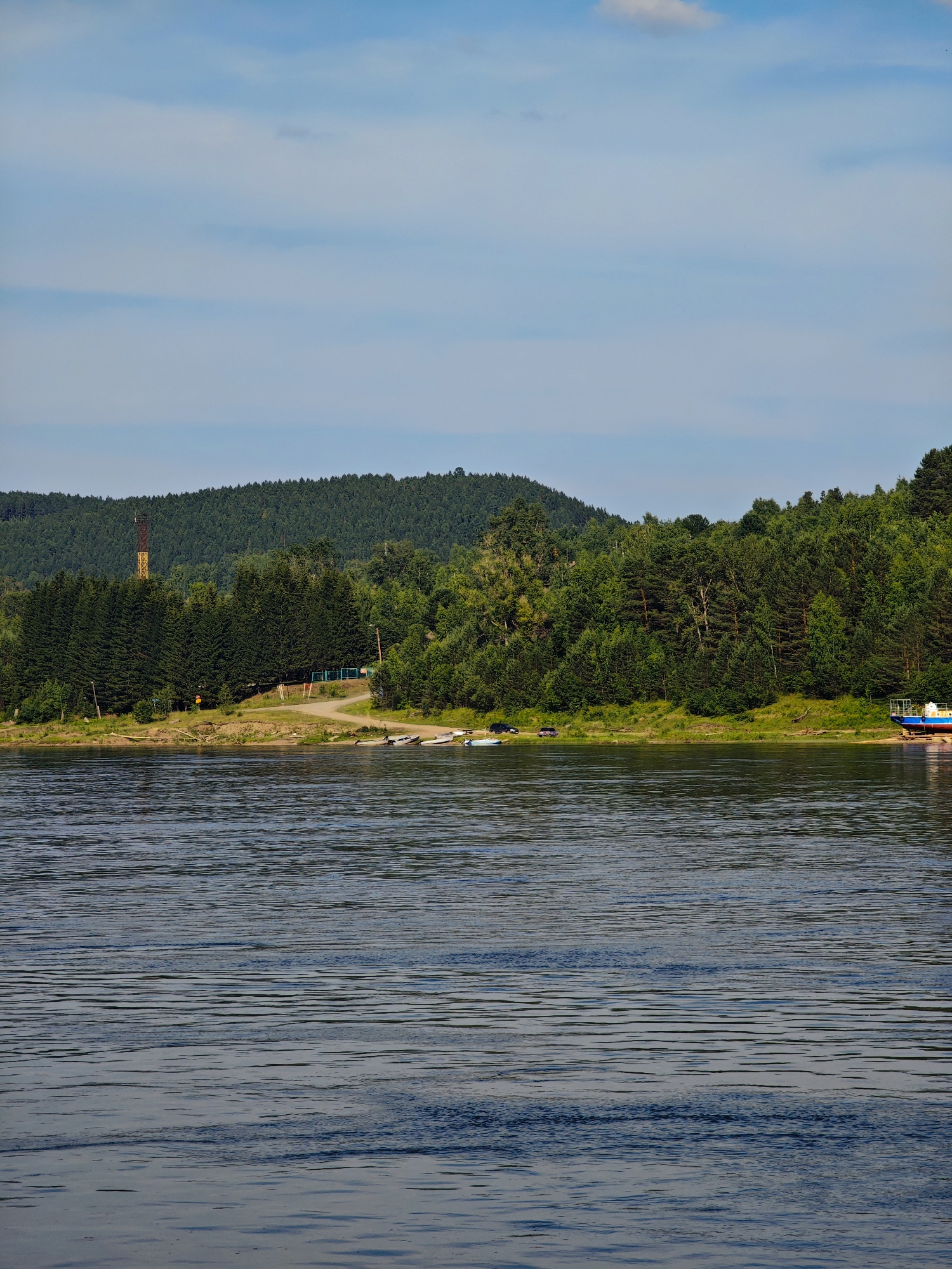 The image shows a wide, calm river with ripples on its surface, reflecting the blue sky and scattered clouds. The riverbank is lined with lush green trees, forming a dense forest that stretches up to a rolling hill. On the left side of the riverbank, a tall, narrow industrial chimney with a yellow and brown striped pattern stands out against the green backdrop. There are several small boats pulled up on the sandy shore near the trees. Further to the right, a larger blue and white boat is partially visible on the shore, possibly undergoing maintenance or repairs. The scene appears to be set in Predivinsk, Russia, a city located on the Yenisei River.