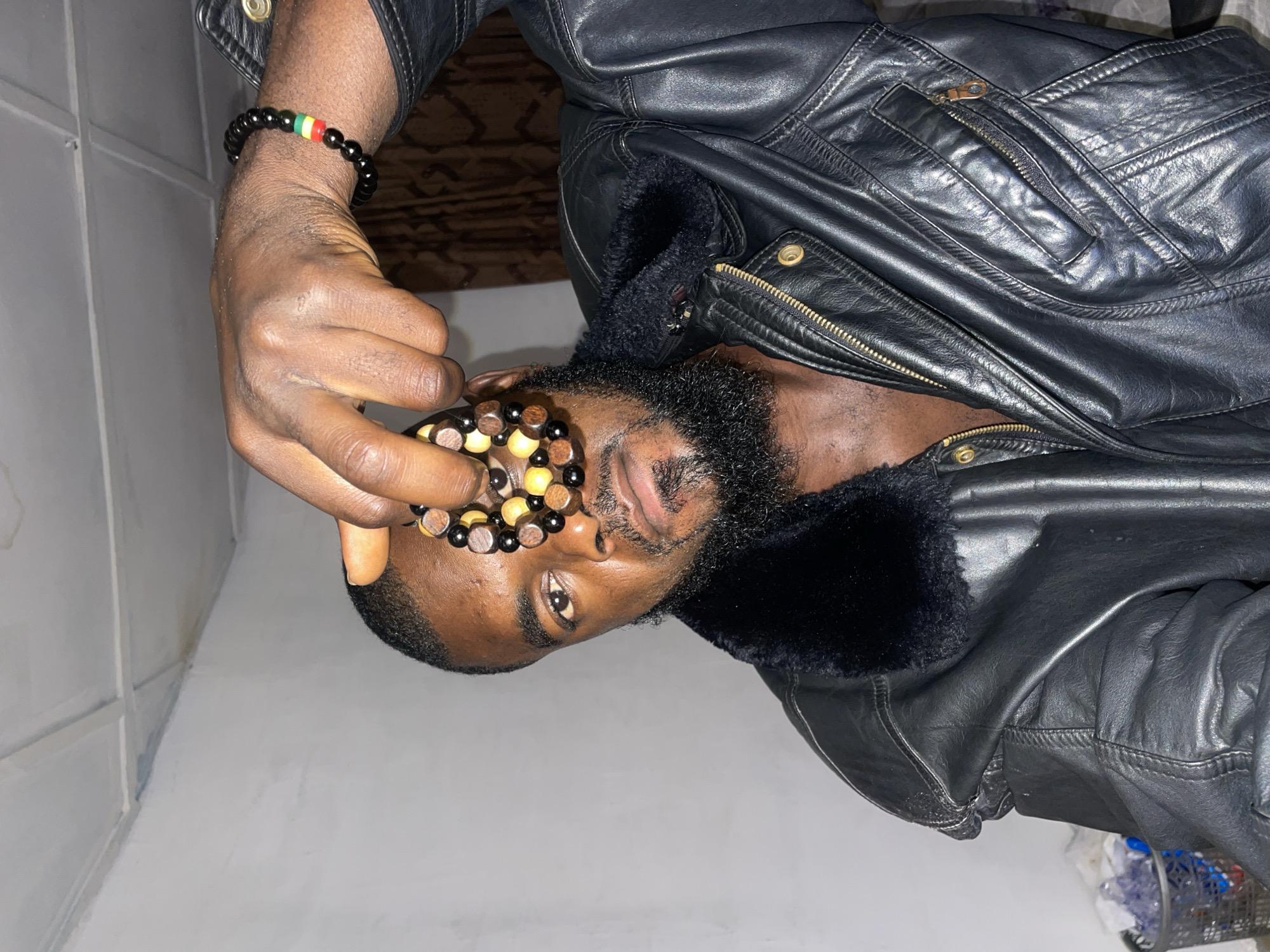 A man with dark skin and a beard is holding a beaded bracelet in front of his face. He is wearing a black leather jacket with a fur collar. He is also wearing another beaded bracelet on his wrist. The background is a white wall and what appears to be a wooden ceiling or wall panel. The location is unknown, as there is no information in the image to suggest it is in Jos, Nigeria.