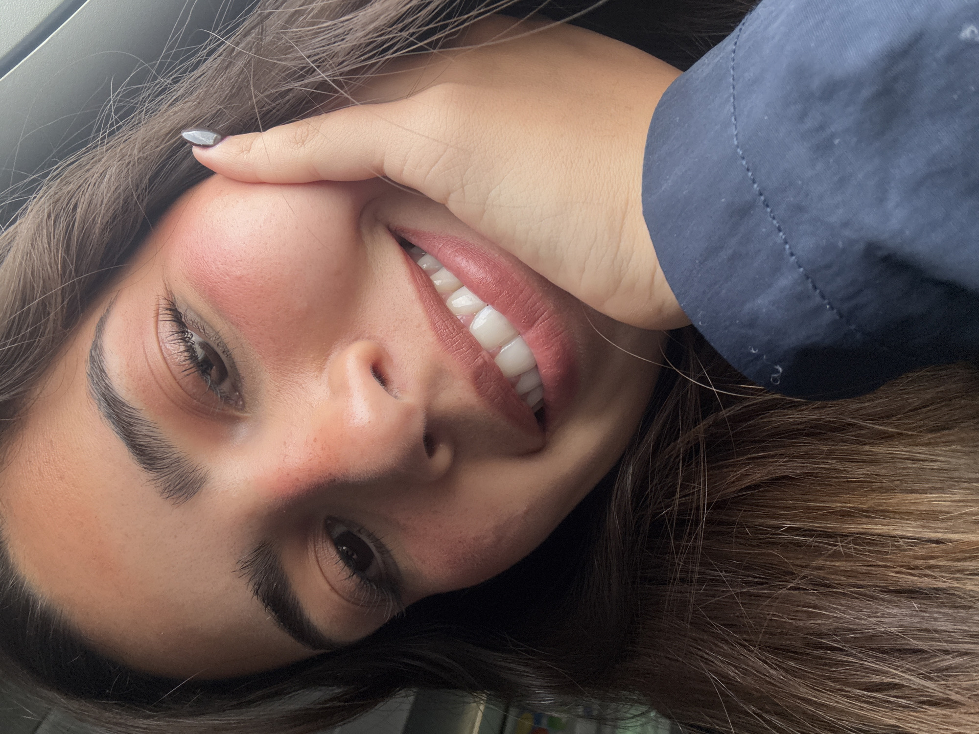 A close-up, rotated portrait shot depicts a person with light skin, dark eyebrows, and brown hair. The individual is smiling, revealing straight, white teeth. Their hand is positioned against their cheek, with a visible gray nail polish. A dark blue sleeve is also present in the frame. No information in the image allows to deduce a location.