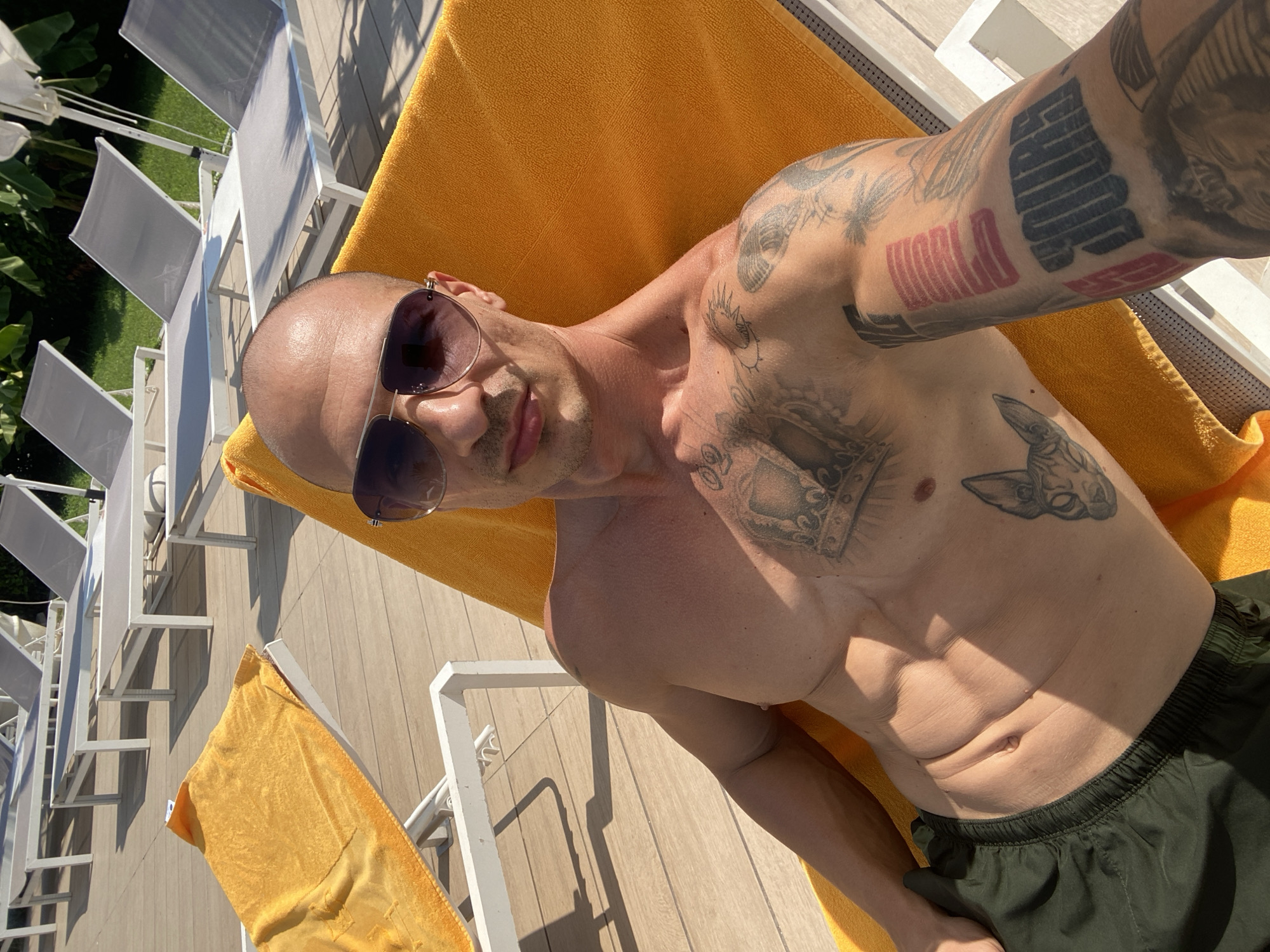 A shirtless man with sunglasses and tattoos is lying on an orange towel on a lounge chair. He is outdoors, with other lounge chairs and greenery visible in the background. The lighting suggests it is daytime. The image does not provide information about the city of Pleven, Bulgaria.
