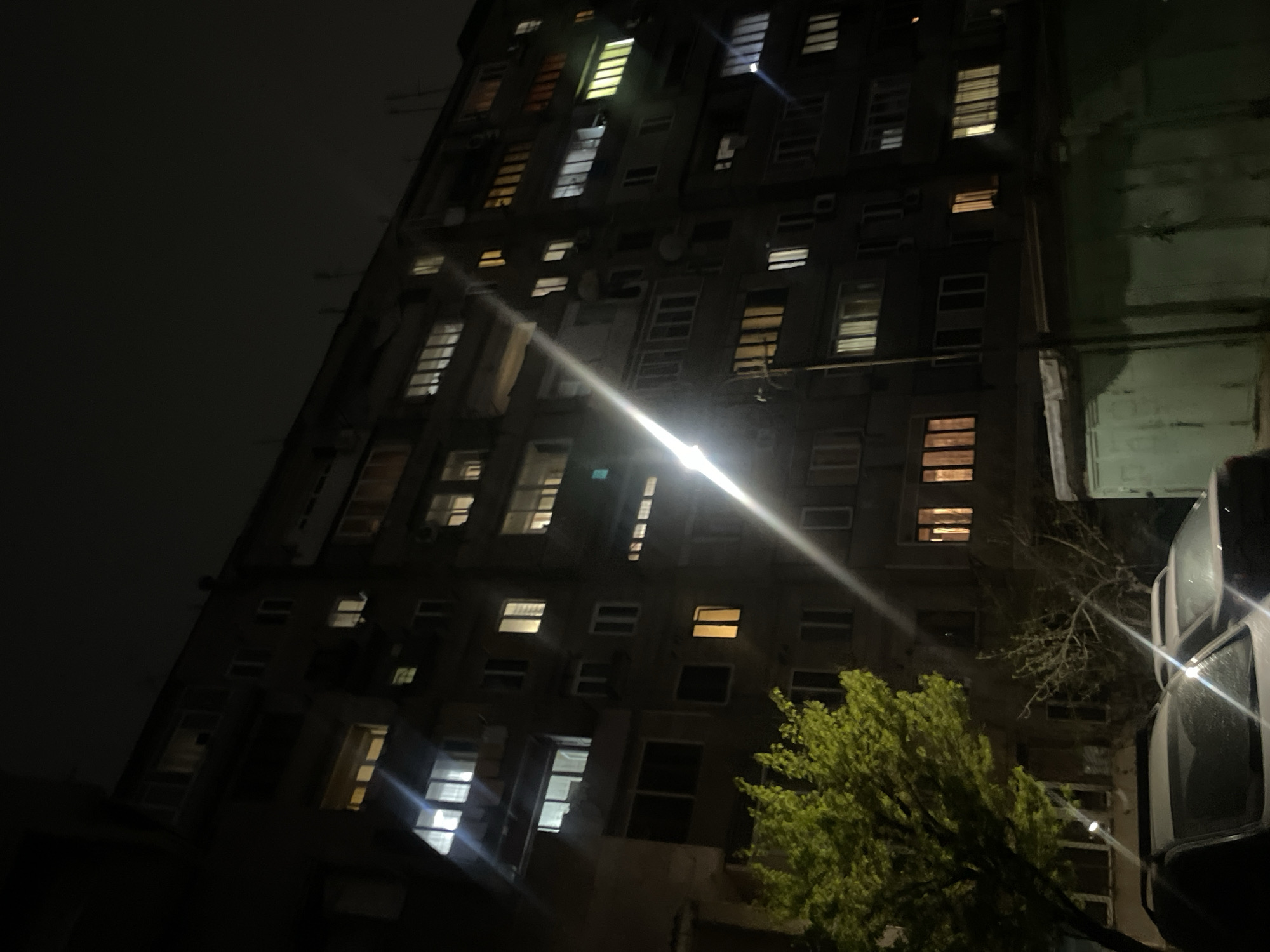 A low-angle shot captures a multi-story building at night. Many windows are visible, some illuminated. A street light shines brightly, causing lens flare. A tree with green leaves is in the foreground, partially obscuring the building. A vehicle is visible on the right, with its windows reflecting light. The sky is dark. While Yerevan, Armenia, cannot be definitively determined from the image, this scene is consistent with an urban environment.