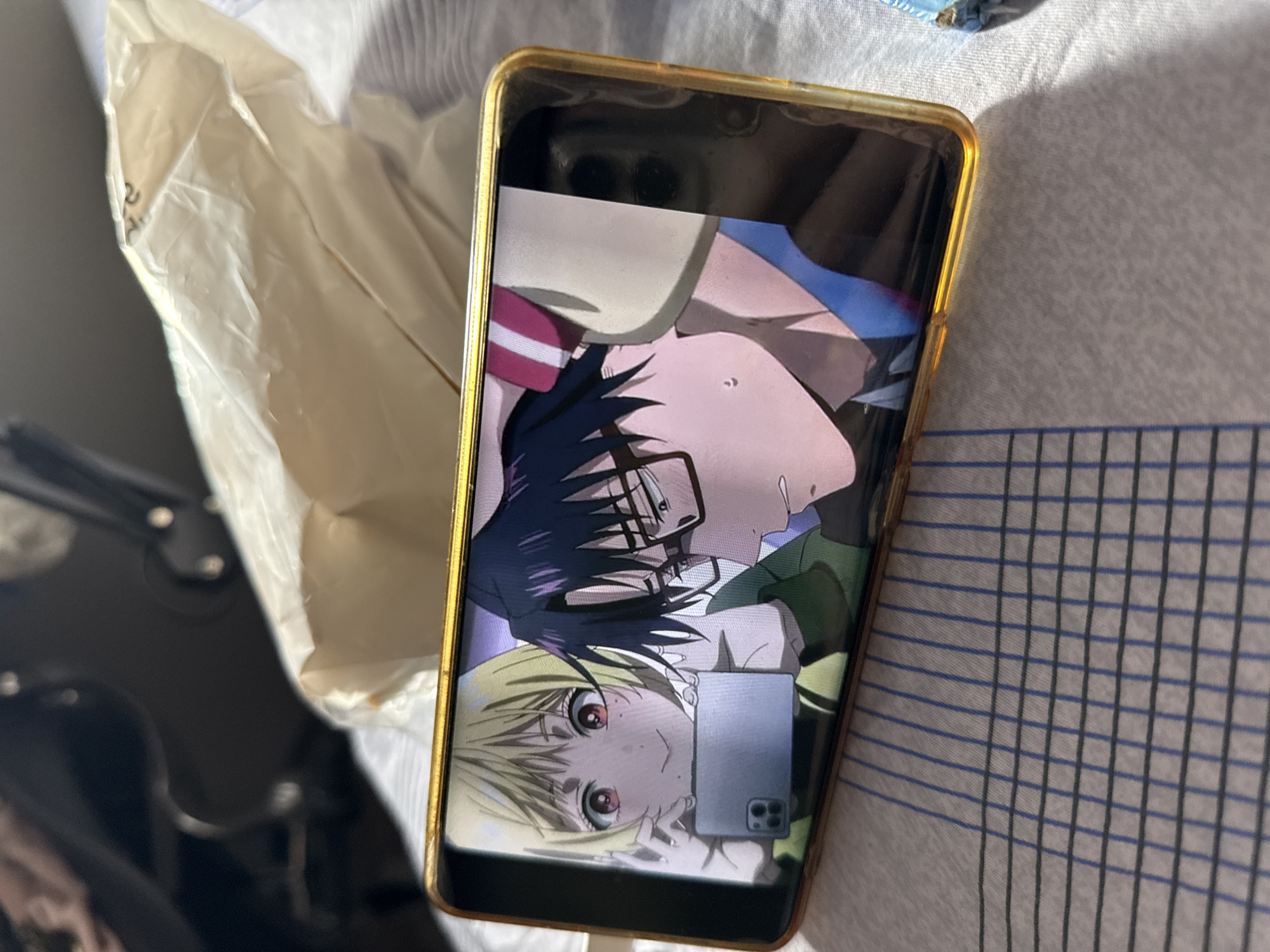 A smartphone with a golden frame is lying on a bed with a blue and gray checkered pattern. The screen of the smartphone displays an anime image featuring a blonde character with red eyes and a male character with black hair and glasses. A white plastic bag is situated to the left of the phone. A black object with silver accents is visible in the lower-left corner of the image. There is no indication of Moscow, Russia.