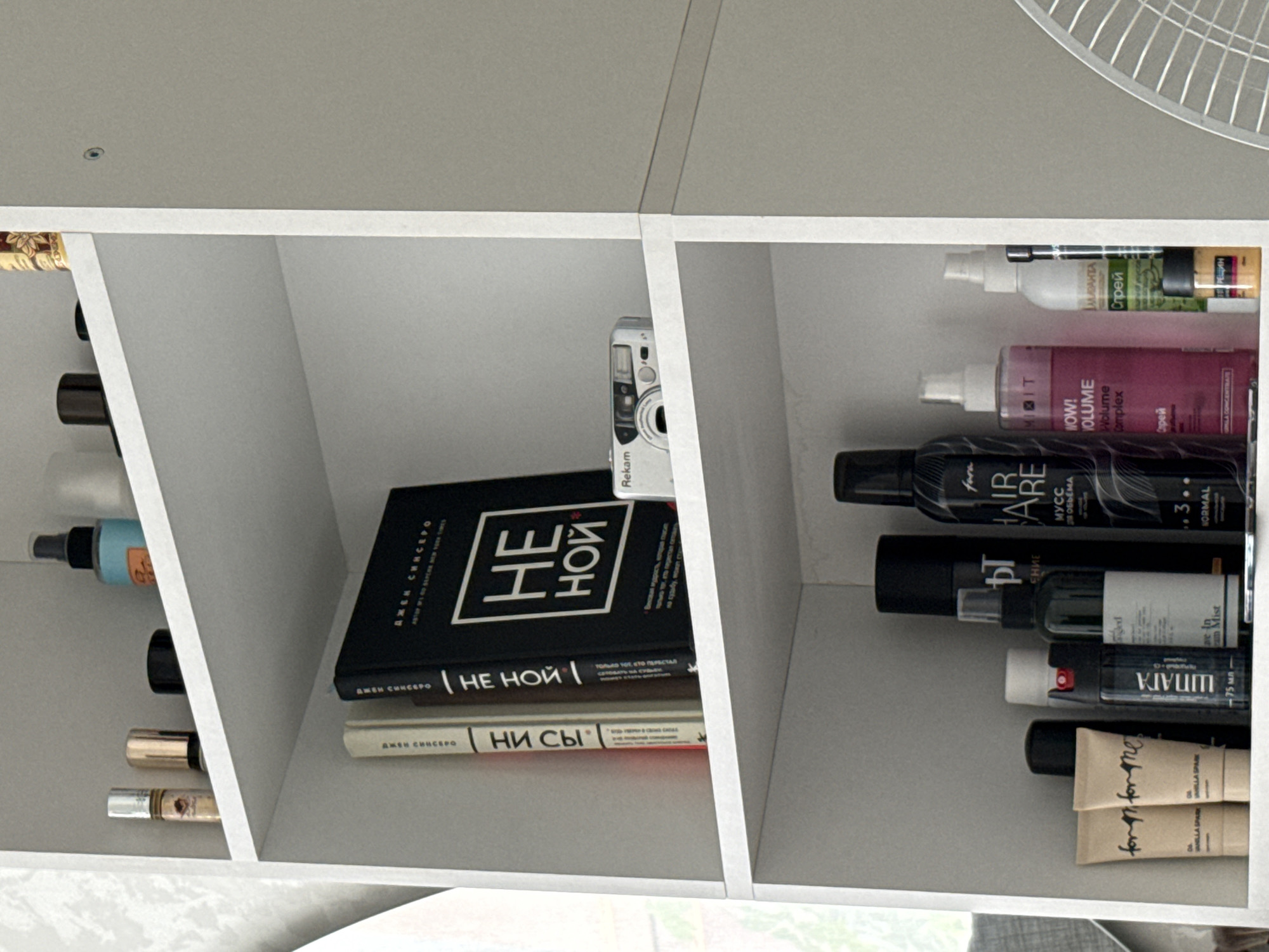 The image depicts a white, multi-tiered shelf unit filled with various items. The top shelf holds several cosmetic and personal care products, including small bottles and jars, as well as a black container. A blue spray bottle with a gold-colored label is also visible. The second shelf down features two books standing upright, one with a black cover and white text, and another with a lighter spine. Next to the books is a small silver digital camera with a lens. The third shelf is densely packed with an assortment of cosmetic products, including tubes, bottles, and spray cans, in various sizes and colors, some with visible labels in Cyrillic. A small fan is partially visible on the right side of the image, suggesting it is in a room. There are no people or any indications of the city of Червишево, Russia.
