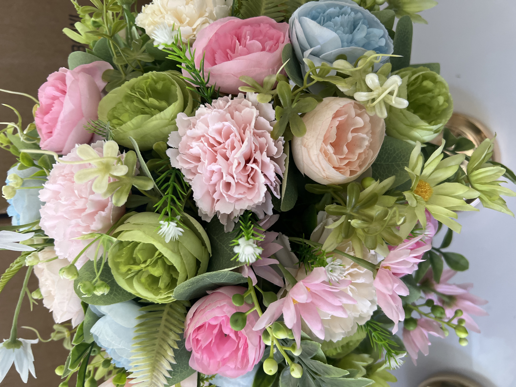 This is a close-up, eye-level, medium shot of an artificial floral arrangement. The bouquet is predominantly composed of pink, pale pink, light blue, cream, light green, and pale orange flowers, interspersed with artificial green foliage. Some flowers are in full bloom with many petals, while others are tighter buds. Small white flowers with sparse petals and green stems are also present. The arrangement is in a gold vase, the rim of which is partially visible at the bottom right of the frame. The background is blurred, with the upper portion appearing to be a dark brown, possibly cardboard, surface, and the lower portion a white surface. There are no people or animals in the image. There is no information that can identify the location as Eshanguzar, Uzbekistan.