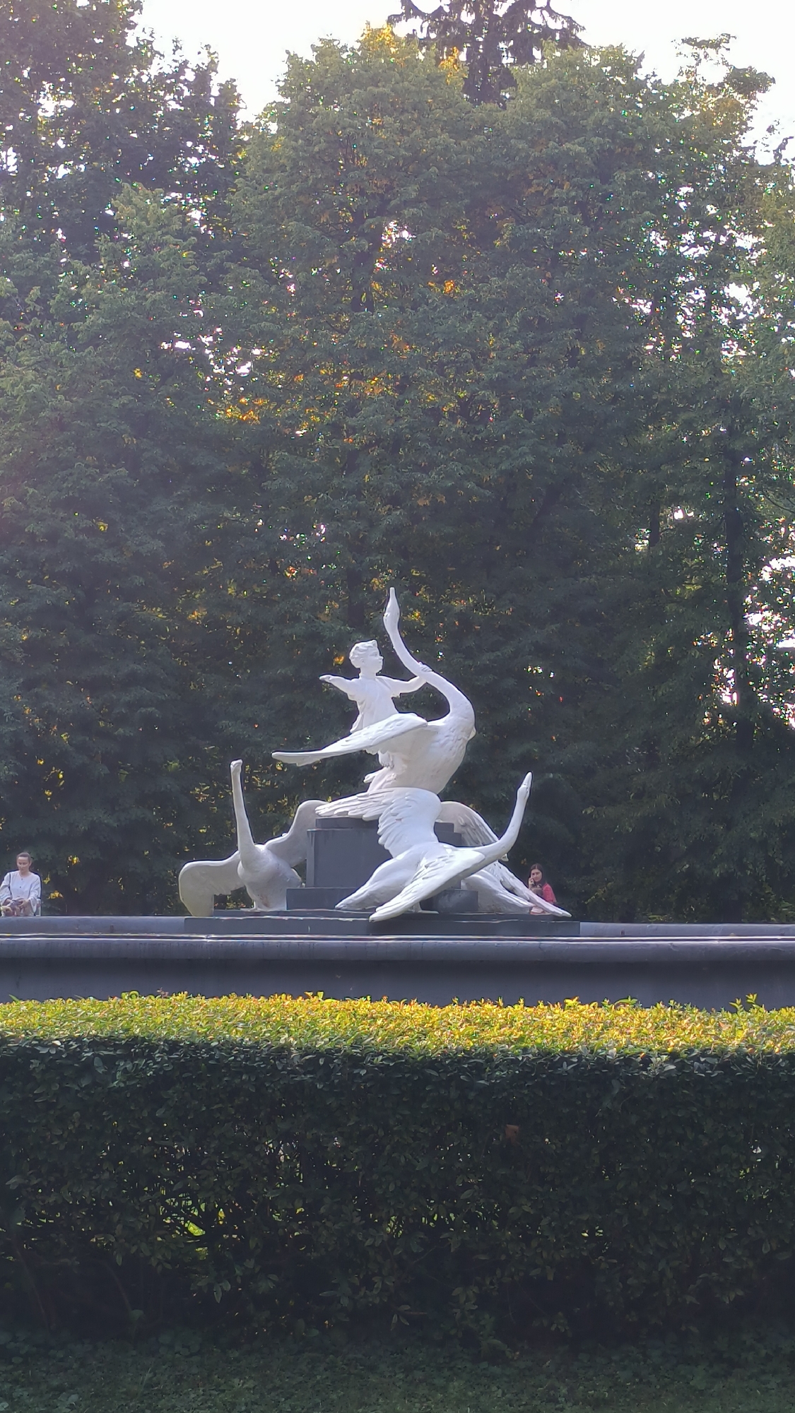 The image shows a white sculpture of a child riding on the backs of swans, set against a backdrop of dense green trees. The sculpture is located in the city of Lviv, Ukraine, and appears to be part of a fountain or a monument. In the foreground, there is a dark grey base, topped with a neatly trimmed green hedge. Two individuals are partially visible: one person is standing to the left of the sculpture, and another person is seated to the right. The lighting suggests it is daytime, with sunlight filtering through the trees, creating dappled patterns. The overall scene is serene and picturesque.