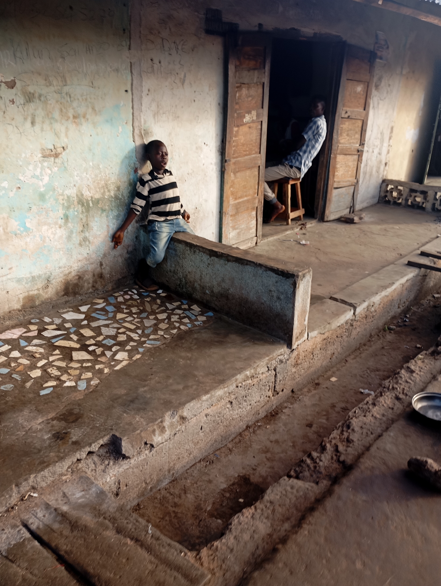 In Shendam, Nigeria, a young boy wearing a striped shirt and jeans leans against a distressed, blue-and-white painted wall covered in graffiti. He is positioned on a concrete platform with a mosaic of broken tiles. To the right, a man in a checkered shirt and light-colored pants sits on a stool inside a doorway. The scene depicts an outdoor area with worn concrete flooring, a drainage ditch, and rough-hewn walls, suggesting a simple or impoverished urban environment.