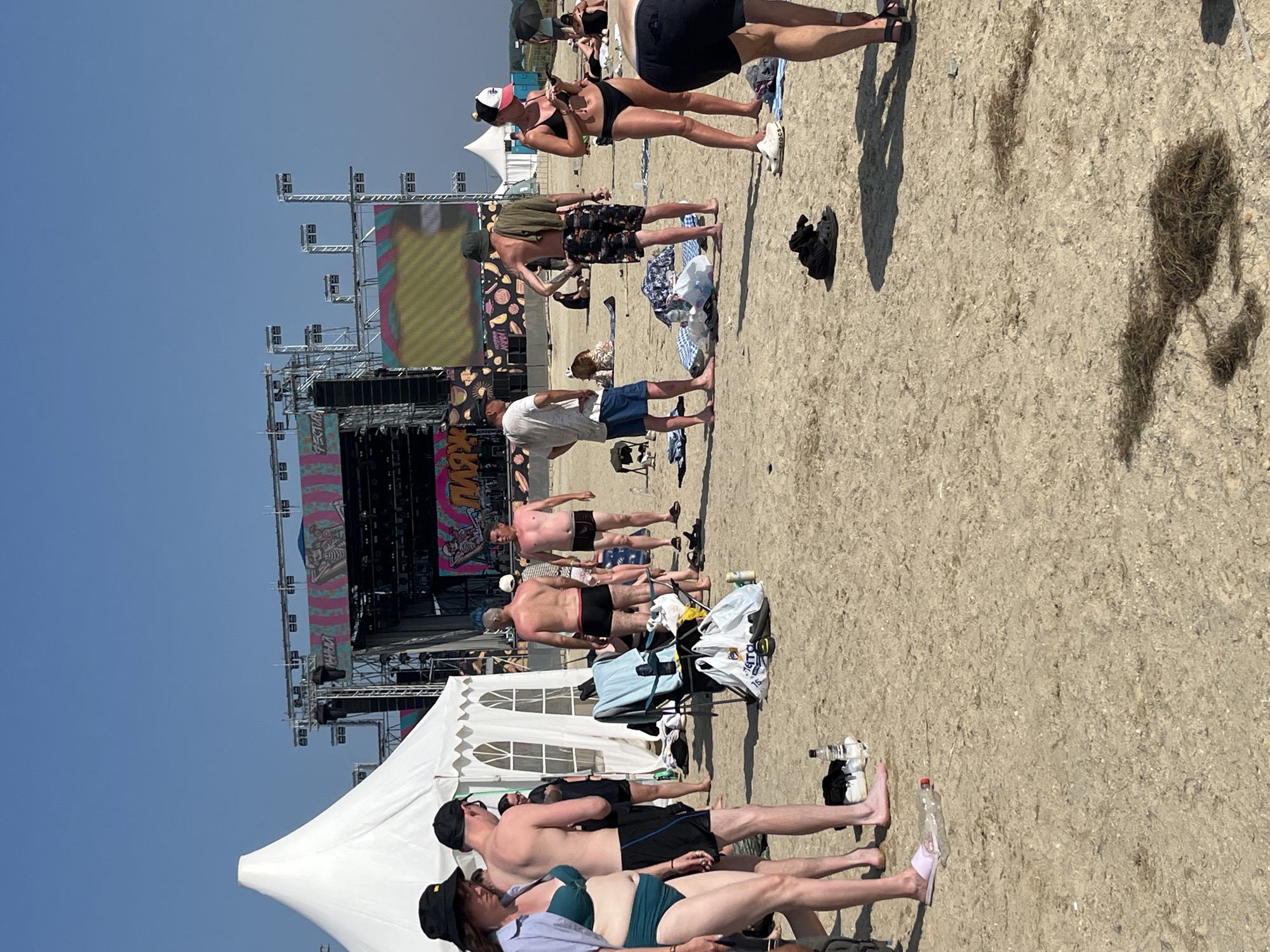 The image depicts a scene at a beach festival in Surakovo, Russia. A large stage with multiple screens and lighting rigs is visible in the background, suggesting a musical performance or event is underway. White tents are set up on the sand, likely for vendors or shelter. Numerous people are scattered across the beach, dressed in casual summer attire, including swimwear. Some individuals are standing and talking, while others appear to be relaxing or preparing to watch the event. Beach items such as bags, towels, and chairs are present on the sand. The environment is sandy with sparse vegetation and a clear blue sky.
