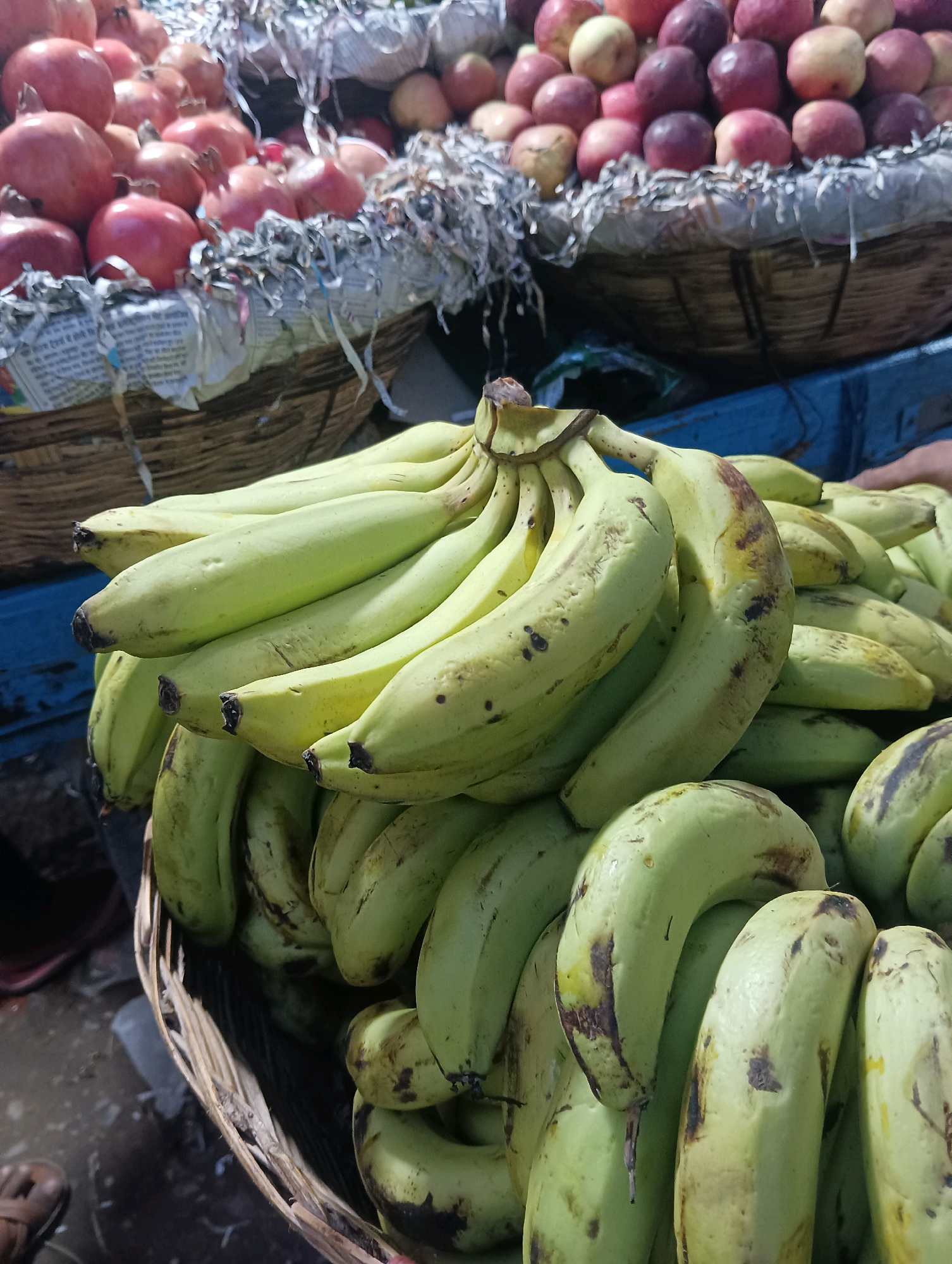 In a market stall in Dakhin Parsa, India, a close-up shot reveals a bounty of fresh fruits. In the foreground, a woven basket overflows with a generous bunch of unripe, green bananas, some showing small brown spots. Behind them, more bananas are piled high, their pale green skins marked with darker blemishes. In the background, other baskets are visible, one filled with vibrant red pomegranates and another with a mix of red and yellow apples. The fruits appear to be for sale, suggesting a lively commercial environment.
