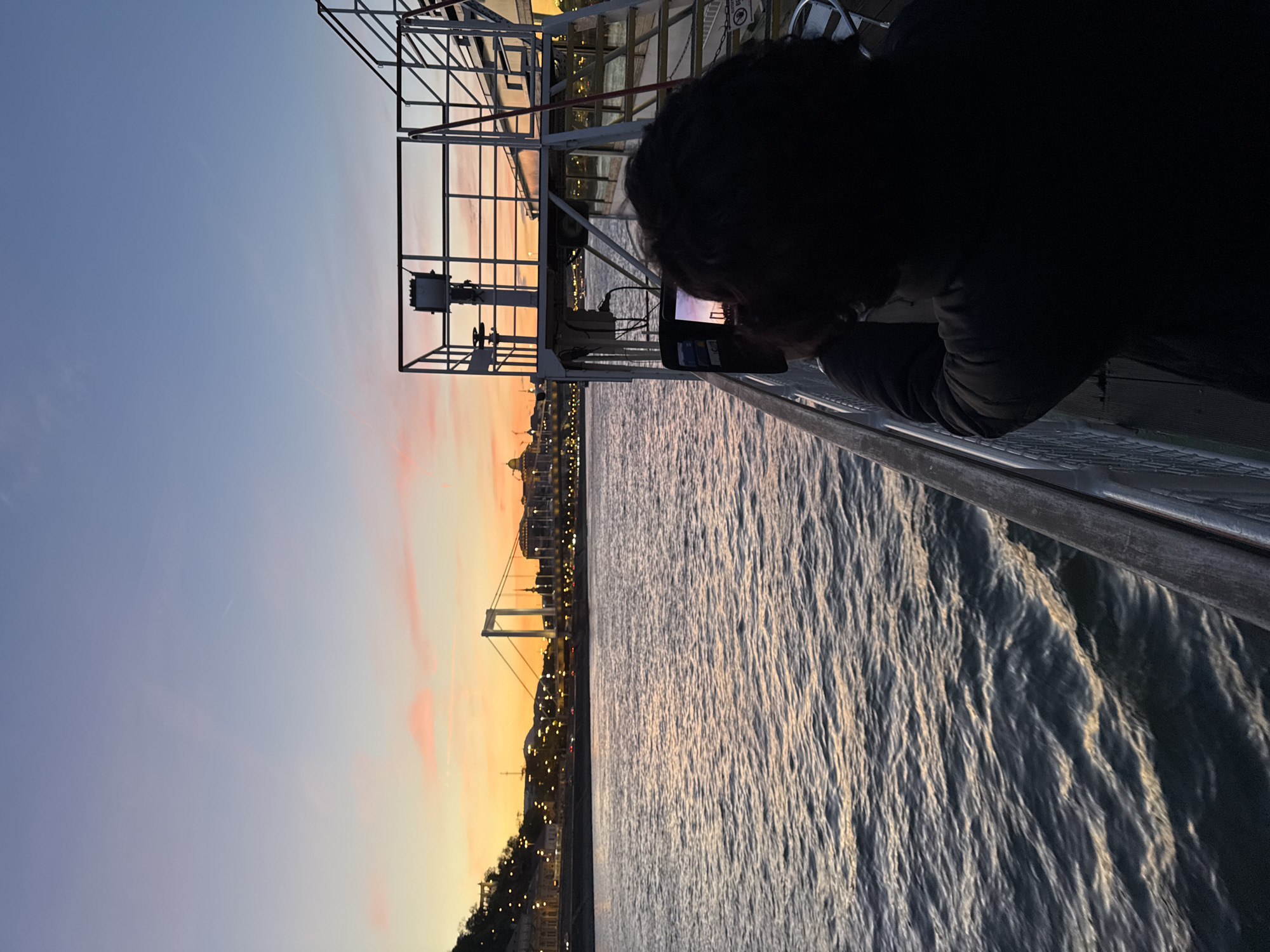 The image captures a scenic twilight view over a body of water in Budapest, Hungary. The primary subject is a person, seen from behind with dark, curly hair, leaning against a metal railing or structure (possibly part of a boat or pier). They are holding a smartphone horizontally, appearing to photograph or video record the illuminated cityscape across the water.
The scene is dominated by the expansive river, likely the Danube, with its surface showing ripples reflecting the fading light. On the far bank, the city skyline is visible, brightly lit with numerous artificial lights, indicating late evening or early night. A prominent suspension bridge with white towers, resembling the Elisabeth Bridge, stretches across the river, its structure outlined by lights. Behind the city, the sky transitions from a soft blue at the top to vibrant orange and pink hues towards the horizon, characteristic of a sunset or twilight.
The person's dark silhouette contrasts with the brighter background of the city lights and colorful sky. The metal framework they are leaning on occupies the right side of the image, adding a foreground element and suggesting the viewpoint is from a vessel or an elevated platform near the water's edge. No discernible text is visible in the image. The atmosphere is calm and serene, highlighting the beauty of the city at dusk.