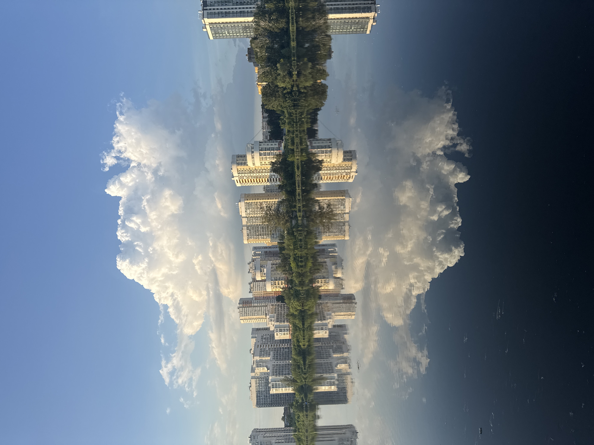 This media file captures a surreal reflection of a city skyline and sky in calm water, oriented vertically. The scene depicts a linear arrangement of modern apartment buildings with numerous windows, interspersed with a dense line of green trees. These structures and the natural foliage are mirrored perfectly in the water below, creating an illusion of two identical, inverted worlds. A large, fluffy white cloud dominates the upper left portion of the sky, with smaller clouds scattered throughout. The sky transitions from a bright blue on the left to a darker, almost black hue on the right, possibly indicating a time around sunset or sunrise, or a change in atmospheric conditions. The water's surface is exceptionally still, allowing for a sharp and clear reflection, broken only by subtle ripples. This suggests a tranquil environment and good weather conditions. The overall impression is one of serene symmetry and the captivating interplay between the built environment, nature, and the reflective quality of water. The specific location context of Yekaterinburg, Russia, can be inferred from the architecture and the visual representation of a city by a body of water. There is no visible text in the image.