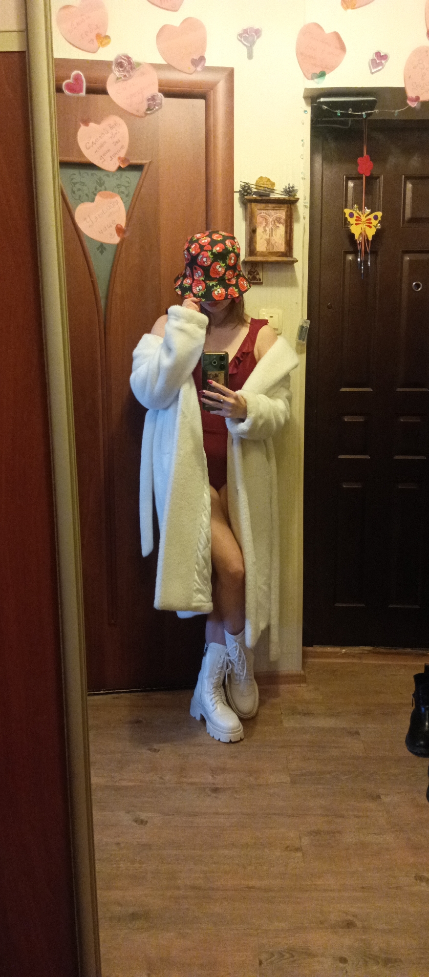 A person is standing in front of a mirror, taking a selfie with a smartphone. The person is wearing a red swimsuit, a white fur coat, a bucket hat with a strawberry pattern, and white lace-up boots with thick soles. The background includes two wooden doors, one with heart-shaped decorations and the other with a butterfly ornament. There is also a small wooden box with a picture on the wall. The scene appears to be indoors, possibly in a hallway. The location is Unknown, Unknown.