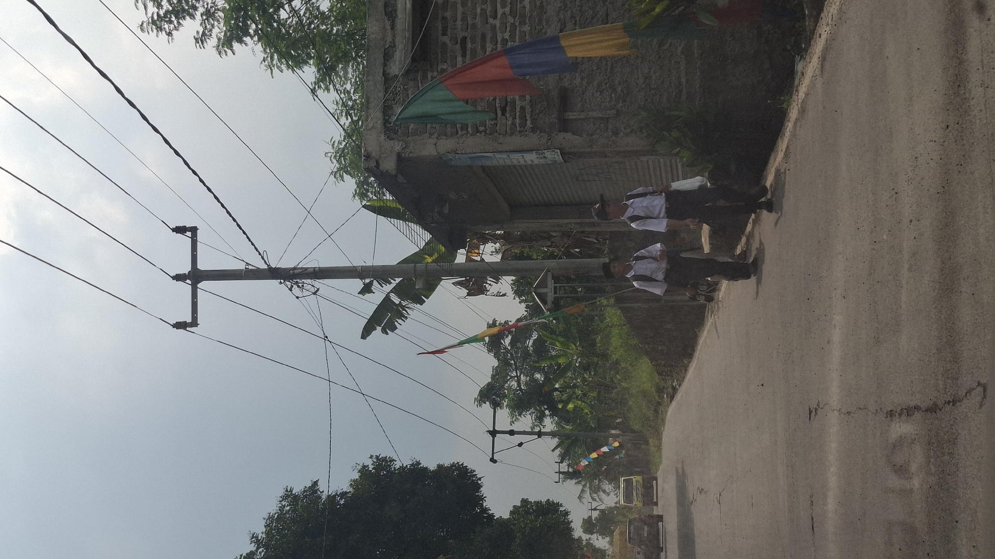 The image depicts a street scene in Sirnagalih, Indonesia, on a cloudy day. Two young males, dressed in school uniforms consisting of white shirts and dark trousers, walk along the right side of a paved road. They are carrying backpacks, suggesting they are returning from or going to school. The road is bordered by a low stone wall on the right and a slightly elevated area with vegetation on the left. Overhead, a network of electrical wires is supported by utility poles. Several colorful flags, possibly celebratory decorations, are visible along the roadside. In the background, a dilapidated brick building with a closed roller door stands on the right, and on the left, vehicles, including a yellow truck and a car, are driving on the road. Lush green trees and banana plants are scattered throughout the environment, indicating a semi-rural or suburban setting. The sky is overcast with bright clouds.