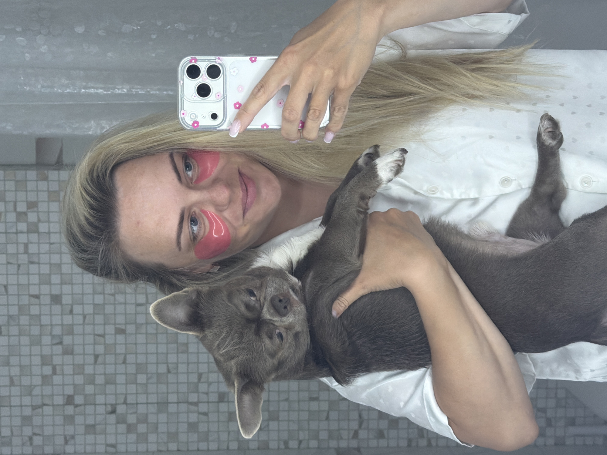 An individual is captured in a mirror selfie with their pet dog. The image is oriented with the woman's head on the left and her body extending horizontally to the right. She has long, blonde hair and is wearing a white collared shirt adorned with subtle grey heart patterns. Pink, gel-like under-eye patches are visible on her face, indicating a beauty or relaxation routine. She smiles gently, looking towards the camera.
In her right hand, she holds a white smartphone encased in a clear cover decorated with small pink flower decals. Her nails feature a light pink and white French tip manicure.
Cradled in her left arm and lying on its back is a small, short-haired dog, likely a Chihuahua or similar breed, with greyish-brown fur and a patch of white on its chest. The dog appears calm and relaxed, its paws partially visible and its gaze directed towards the camera.
The background reflects a wall covered in small, light grey square tiles, suggesting an indoor setting, possibly a bathroom or a room with tiled walls. The overall scene conveys a moment of affection and relaxation between the woman and her dog, possibly at her home in Votkinsk, Russia. There is no visible text.
