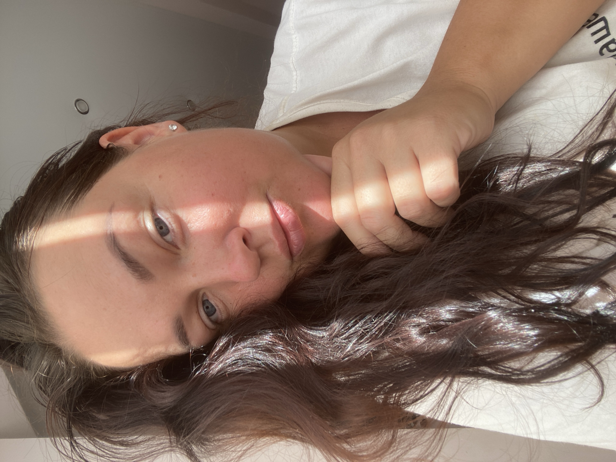 A close-up, waist-up, indoor portrait of a woman is captured in a selfie. She has long, dark brown wavy hair that is partially obscuring her face. Her right hand is resting on her chin. Sunlight casts a diagonal shadow across her face. She is wearing a white t-shirt. The background is blurred, showing a neutral-colored wall and ceiling with light fixtures. The lighting suggests it is daytime, with natural light coming from the side. The woman's facial expression is neutral, and she is looking directly at the camera. There is no identifiable indication of the city of Sochi, Russia within the image.