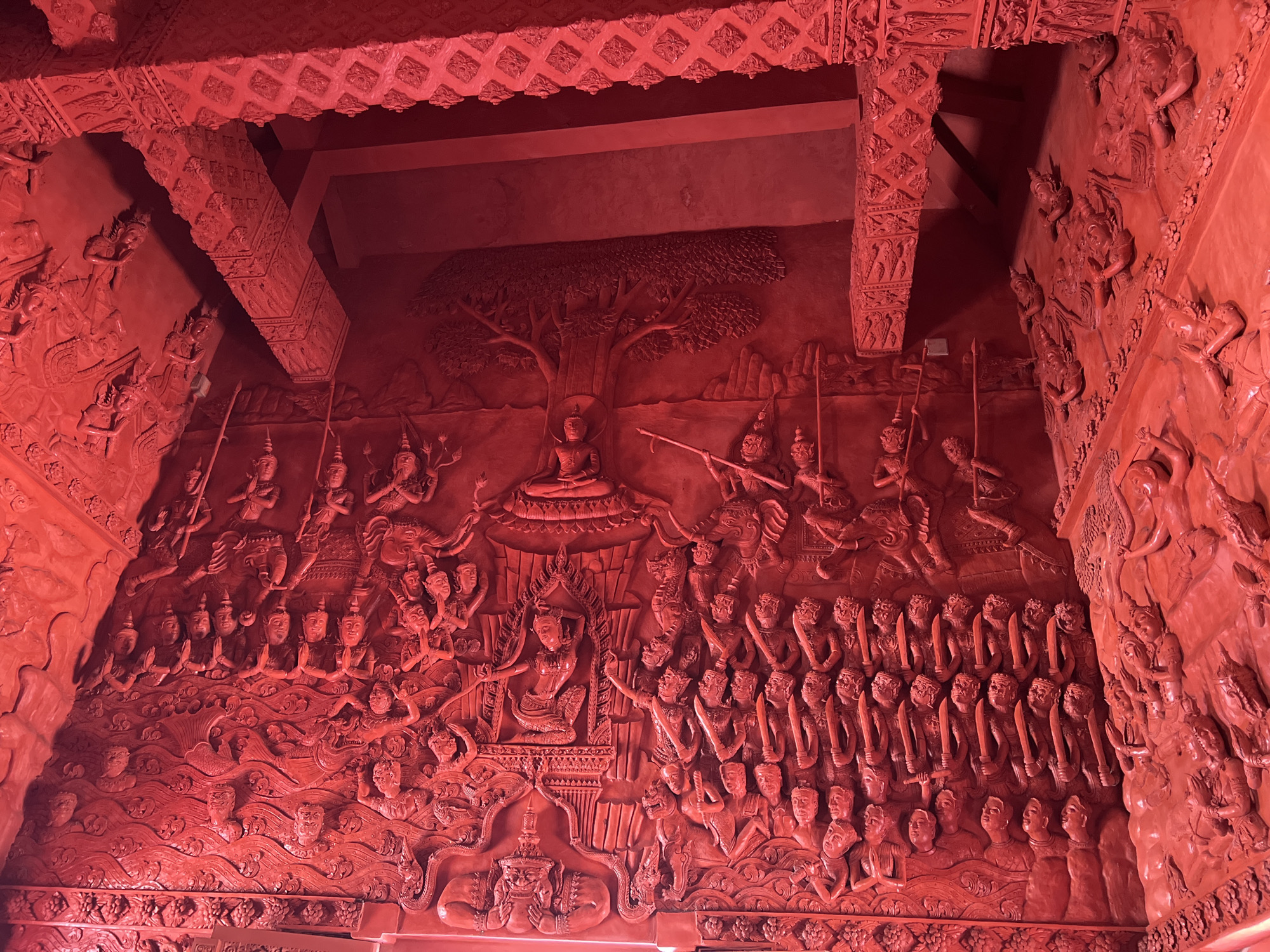 This image shows a highly detailed, entirely red-painted wall carving. The carving depicts a scene with numerous figures, including humans and mythical beings, in various poses and actions. The scene appears to illustrate a narrative or historical event.
In the center of the top portion of the carving, there is a depiction of the Buddha seated under a tree. To either side of the Buddha, figures are holding spears and other weapons.
The lower portion of the carving showcases rows of figures, many of them armed with swords. The figures appear to be arranged in a structured manner, possibly representing an army or procession.
The image is likely from the Red Temple or Wat Sila Ngu, a temple located in Baan Hua Thanon, Thailand. The temple is notable for its striking red color and intricately carved details. The image captures the unique artistic style and cultural significance of the temple.