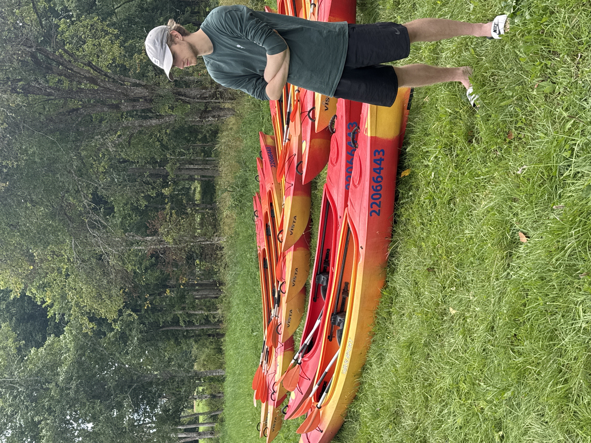 A young Caucasian male wearing a green long-sleeved shirt, black shorts, a grey baseball cap, and white Nike slides stands in a grassy field with his arms crossed. He is looking to his right, away from the camera. In the background, a line of red and yellow kayaks with orange paddles are arranged on the grass. The kayaks have numbers and text printed on them, including "VISTA" and "22066443". Behind the kayaks, a dense line of trees forms the backdrop. The ground is covered in green grass, with some scattered leaves and fallen branches. The lighting suggests an overcast day. The scene appears to be an outdoor recreational area, possibly near a body of water, given the presence of kayaks. The specific location in Vērene, Latvia is not discernible from the image itself.