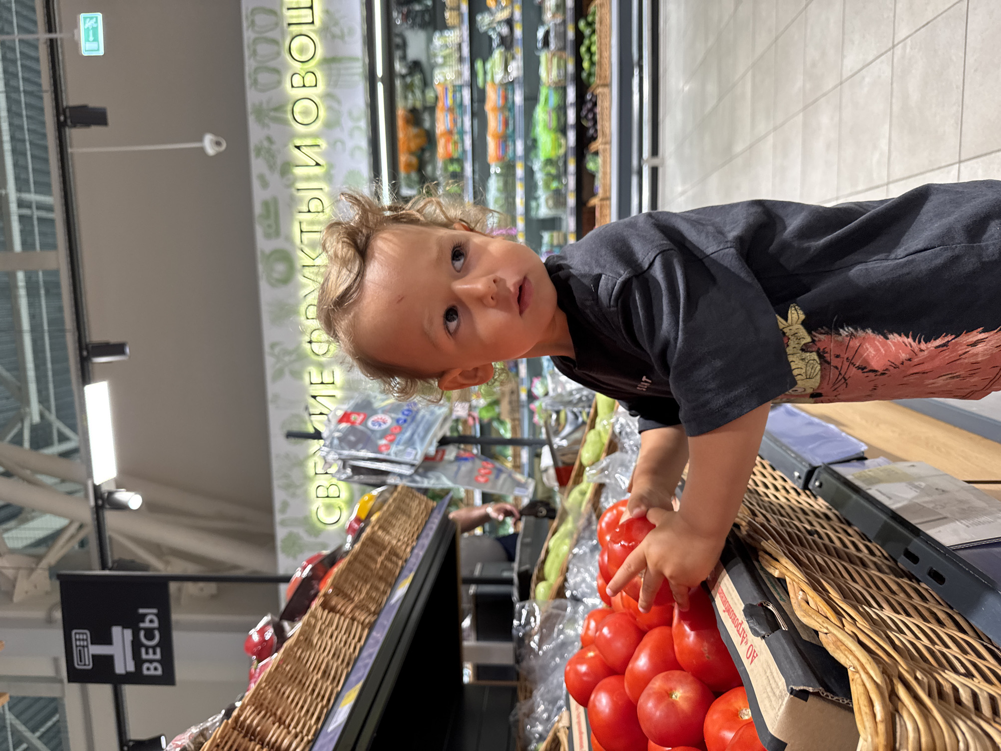 A young child with curly blond hair and brown eyes is standing in a supermarket in Sochi, Russia, reaching for a tomato from a basket of produce. The child is wearing a dark t-shirt with a graphic print. In the background, shelves are stocked with various fruits and vegetables. A sign above the produce display reads "СВЕЖИЕ ФРУКТЫ И ОВОЩИ" (Fresh Fruits and Vegetables). Another sign with an icon of a scale and the word "ВЕСЫ" (Scales) is visible in the upper left corner, indicating a weighing station. The flooring appears to be tiled.