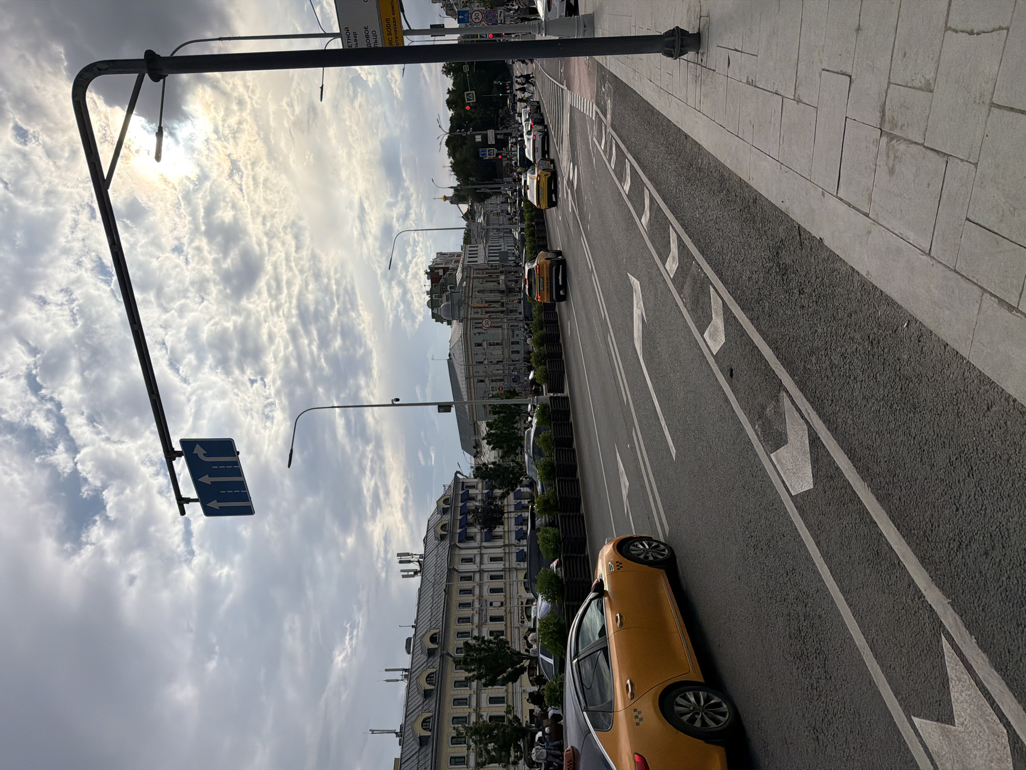 The image shows a city street with multiple lanes of traffic. In the foreground, an orange taxi is visible in the left lane. The street has directional arrows painted on it, indicating that the left lane is for turning left or going straight. Further down the road, several other vehicles, including taxis and cars, are in motion.
On the right side of the street, a sidewalk is present with a lamppost and a traffic sign. In the background, buildings line the street, some with multiple stories and classical architectural features. The sky is partly cloudy with the sun shining through, creating a somewhat overcast atmosphere. The overall scene depicts a bustling urban environment with active traffic flow.