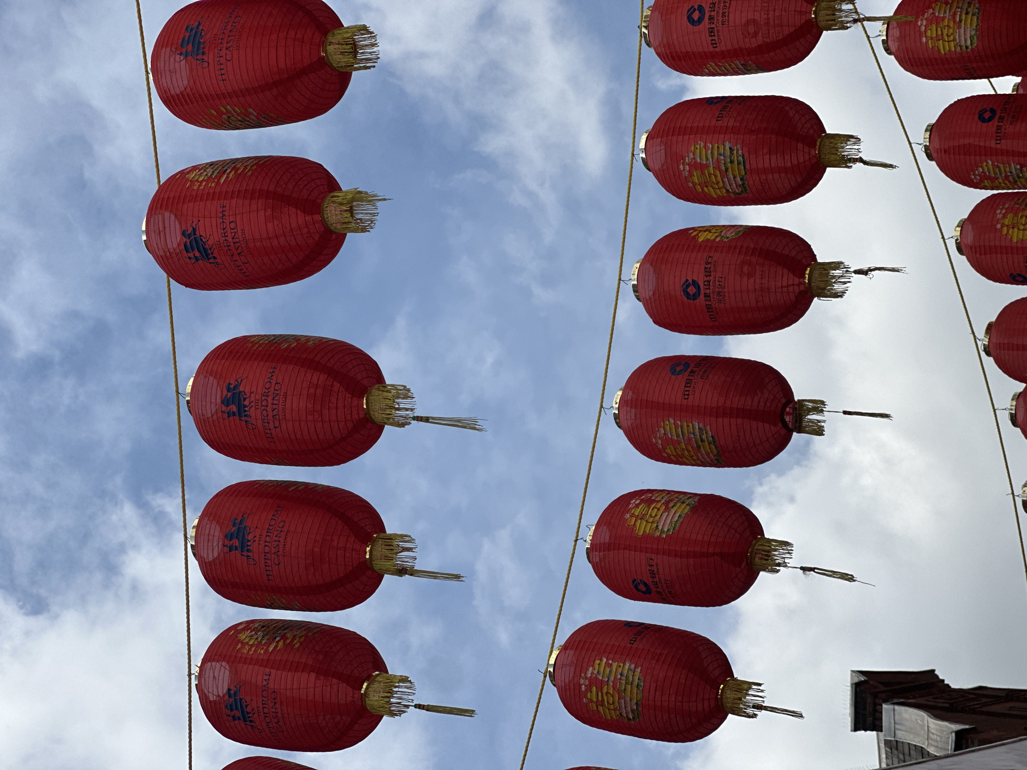 The photograph showcases a vertical arrangement of red Chinese lanterns suspended above a street in Chinatown, London, United Kingdom. The left row of lanterns are branded with "The Hippodrome Casino, London." The right row of lanterns are branded with Chinese writing. The background is filled with a partly cloudy sky. The angle of the shot is from below, looking upward towards the lanterns and the sky. The lanterns are a common decoration in Chinatown, and they create a vibrant, festive atmosphere. In the bottom right, a piece of the surrounding architecture can be seen.