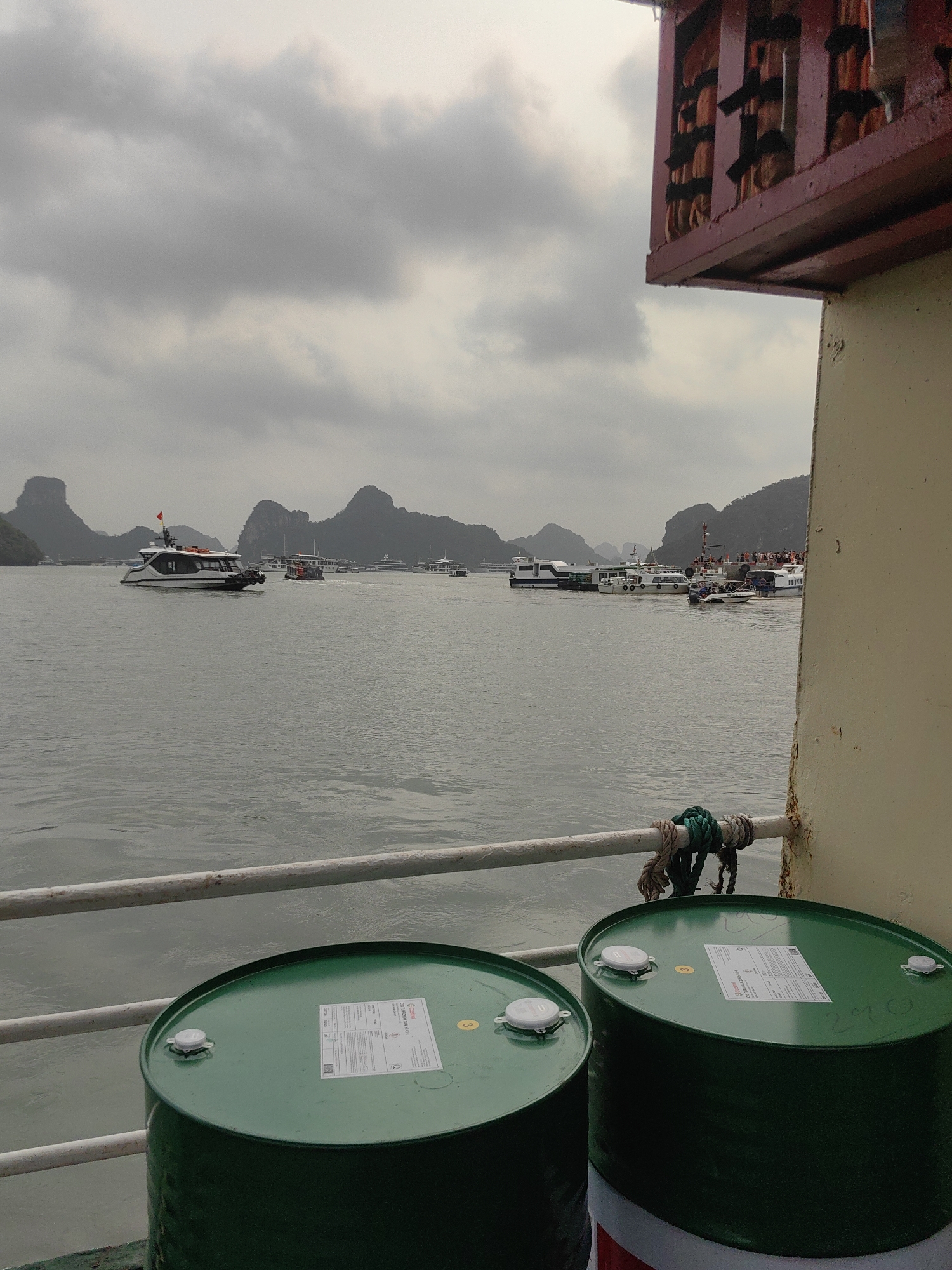 The image shows a cloudy day on the waters of Hải Phòng, Vietnam, with various boats visible in the distance against a backdrop of islands. In the foreground, on what appears to be a boat, are two green drums with white labels, positioned behind a weathered metal railing. Part of the boat's structure is visible on the right side of the image, with peeling paint and a decorative wooden overhang.