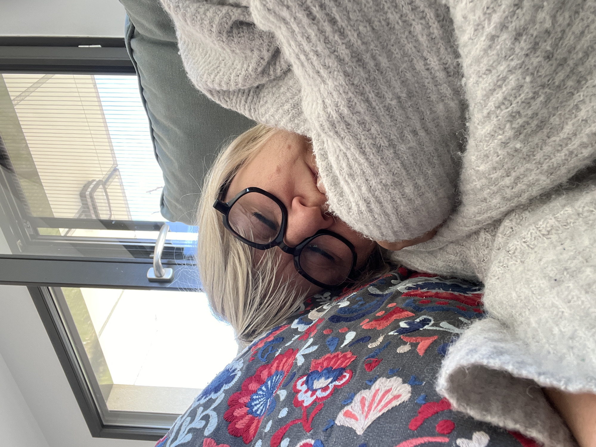 A woman wearing glasses and a gray knit sweater is lying on a colorful pillow. A window with blinds is visible in the background. The location is not specified, and there is no indication that it is in Chambœuf, France.